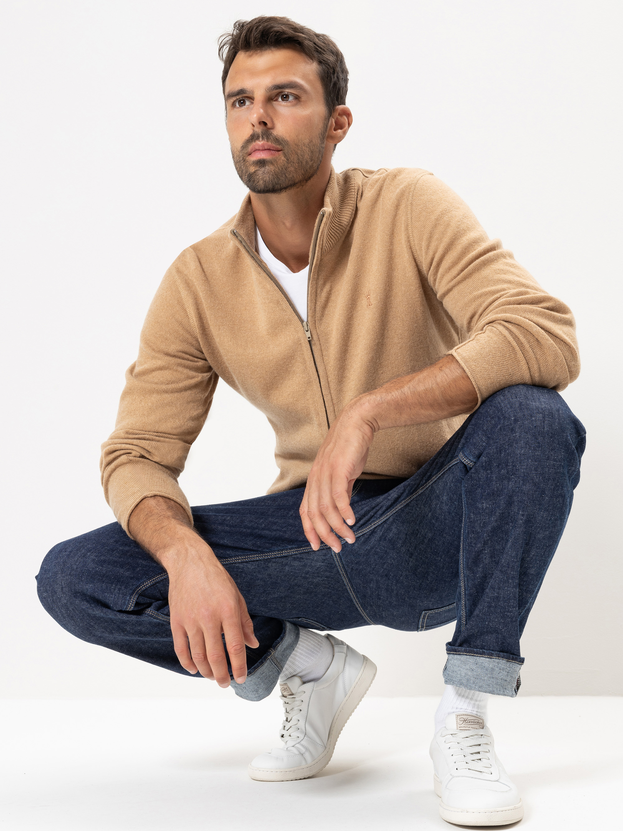 Ein Mann mit kurzen dunklen Haaren und einem Bart, bekleidet mit einem hellbraunen Pullover mit Reißverschluss, einem weißen T-Shirt, hochgekrempelten Bluejeans und weißen Turnschuhen, hockt vor einem schlichten weißen Hintergrund und schaut leicht zu