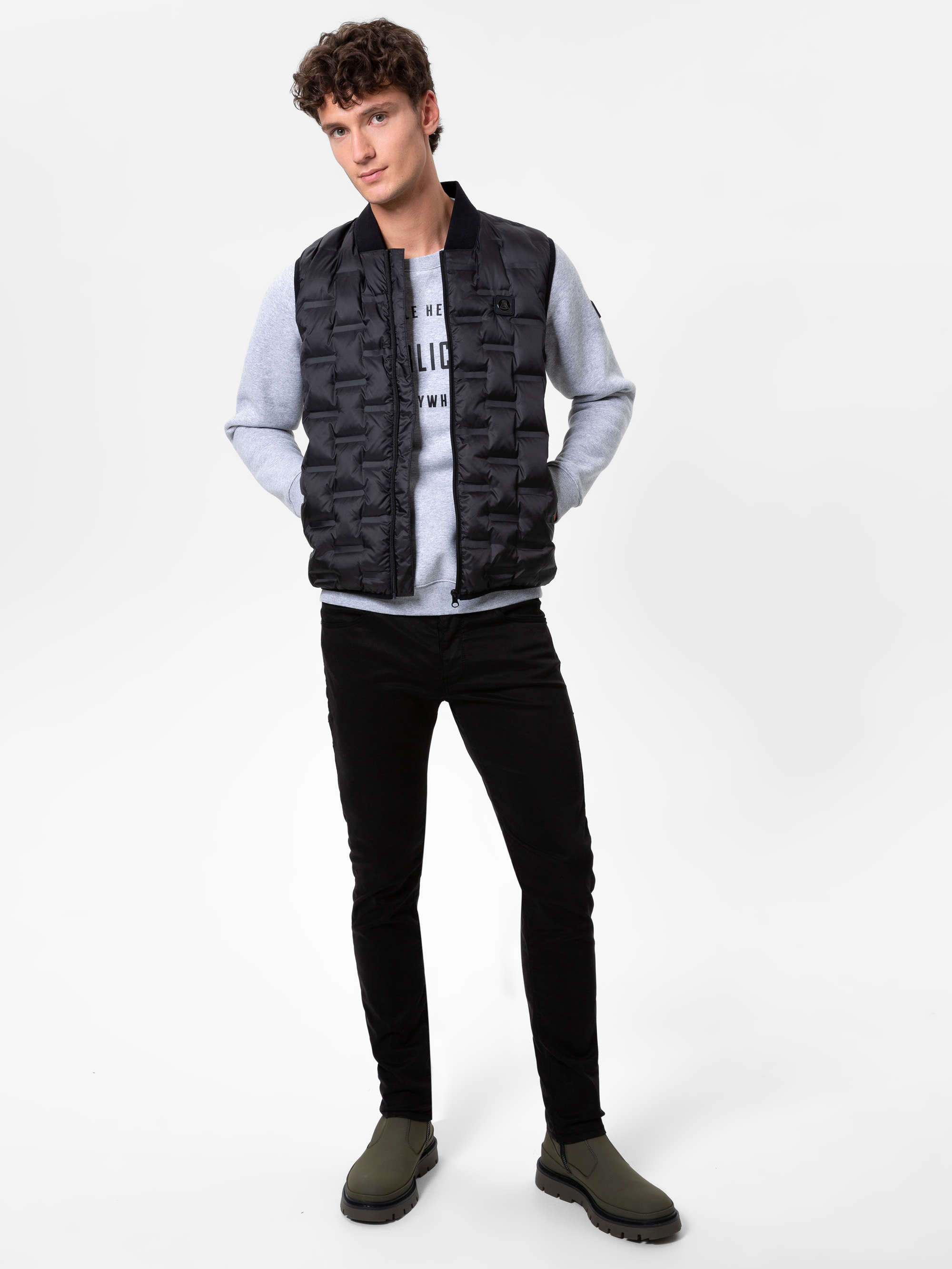 Ein junger Mann mit lockigem braunem Haar posiert vor einem weißen Hintergrund. Er trägt eine schwarze Pufferweste über einem grauen Sweatshirt, schwarze Jeans und olivgrüne Stiefel. Er steht mit einer Hand in der Tasche und schaut in die Kamera.