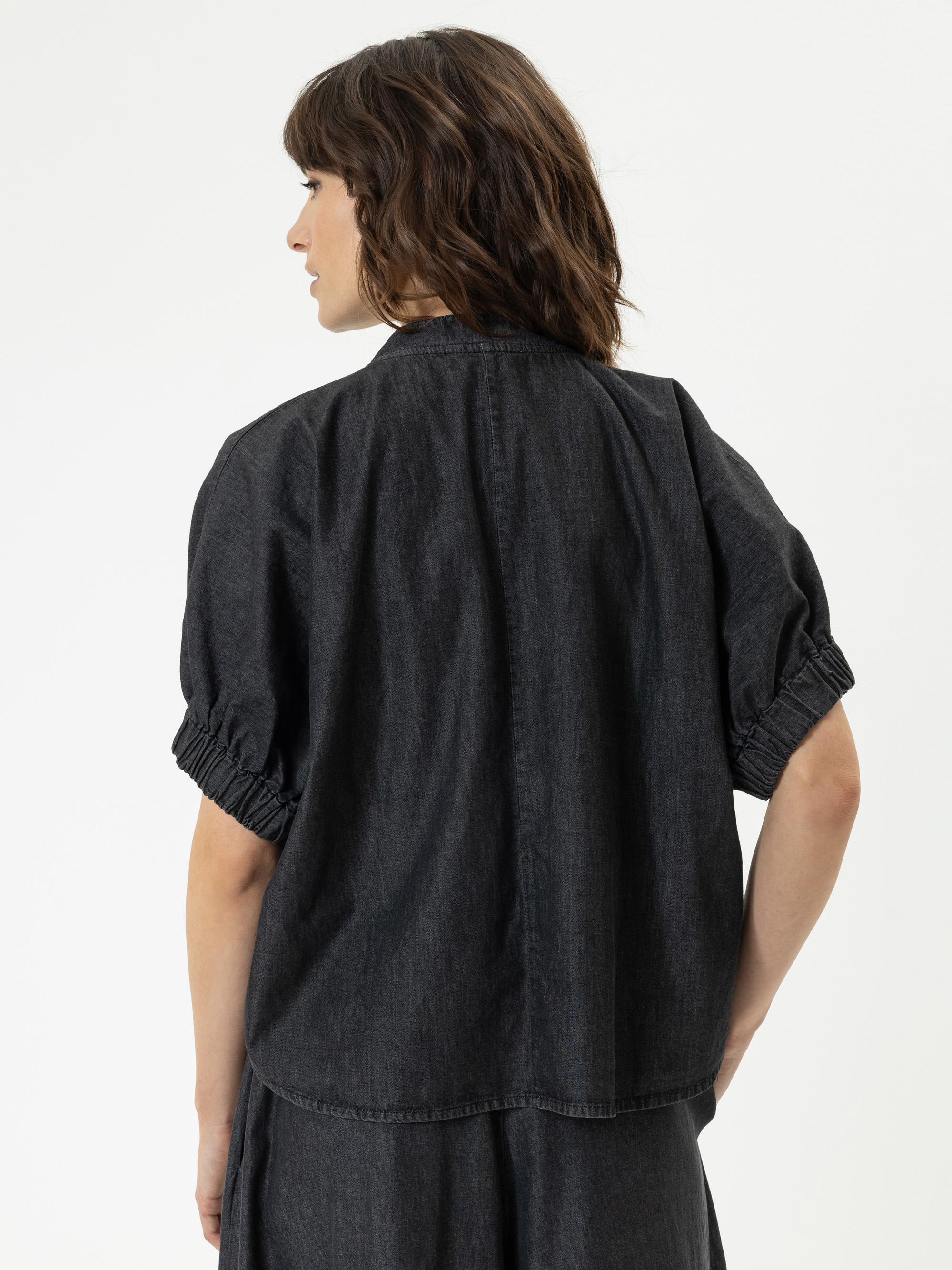 Eine Frau mit schulterlangen braunen Haaren wird von hinten gezeigt. Sie trägt ein locker sitzendes, kurzärmeliges schwarzes Jeansoberteil mit gerafften Ärmeln. Sie steht vor einem schlichten weißen Hintergrund.