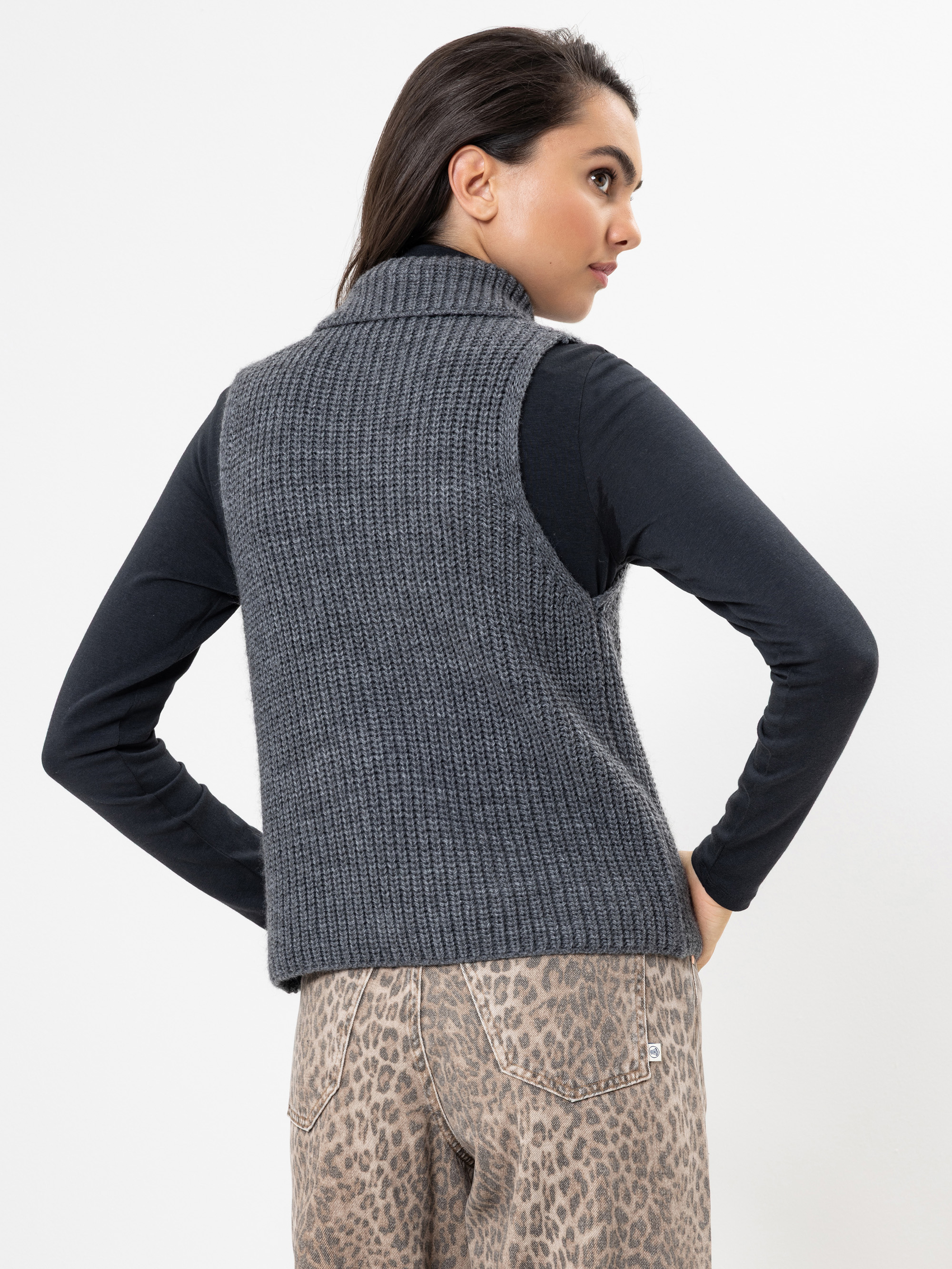 Eine Frau steht mit dem Gesicht nach hinten, trägt eine ärmellose graue Strickweste über einem langärmeligen schwarzen Oberteil und eine hellbraune Hose mit Leopardenmuster, die linke Hand auf die Hüfte gestützt.
