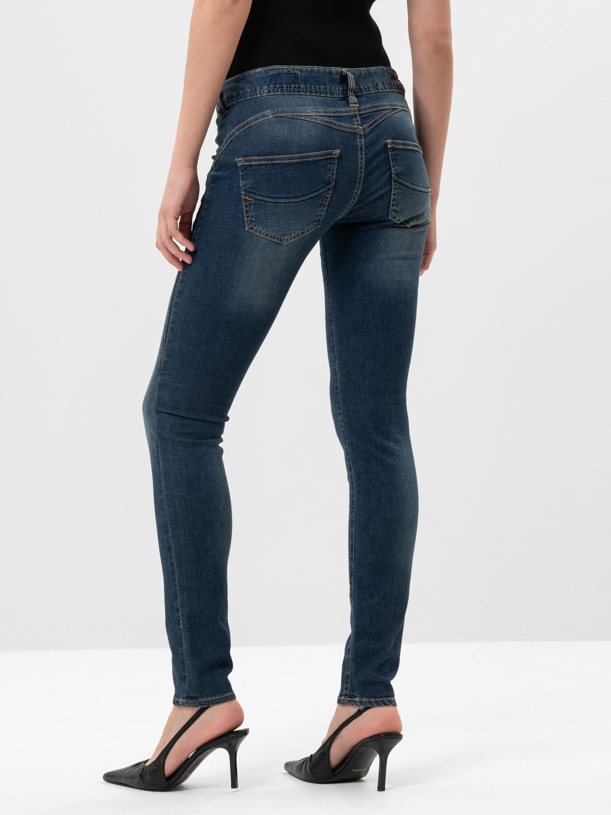 Eine Person, die dunkelblaue Skinny Jeans und schwarze Slingback-Schuhe mit Absatz trägt, steht auf einer weißen Fläche, von hinten gesehen. Die Jeans hat sichtbare Nähte und zwei Gesäßtaschen.