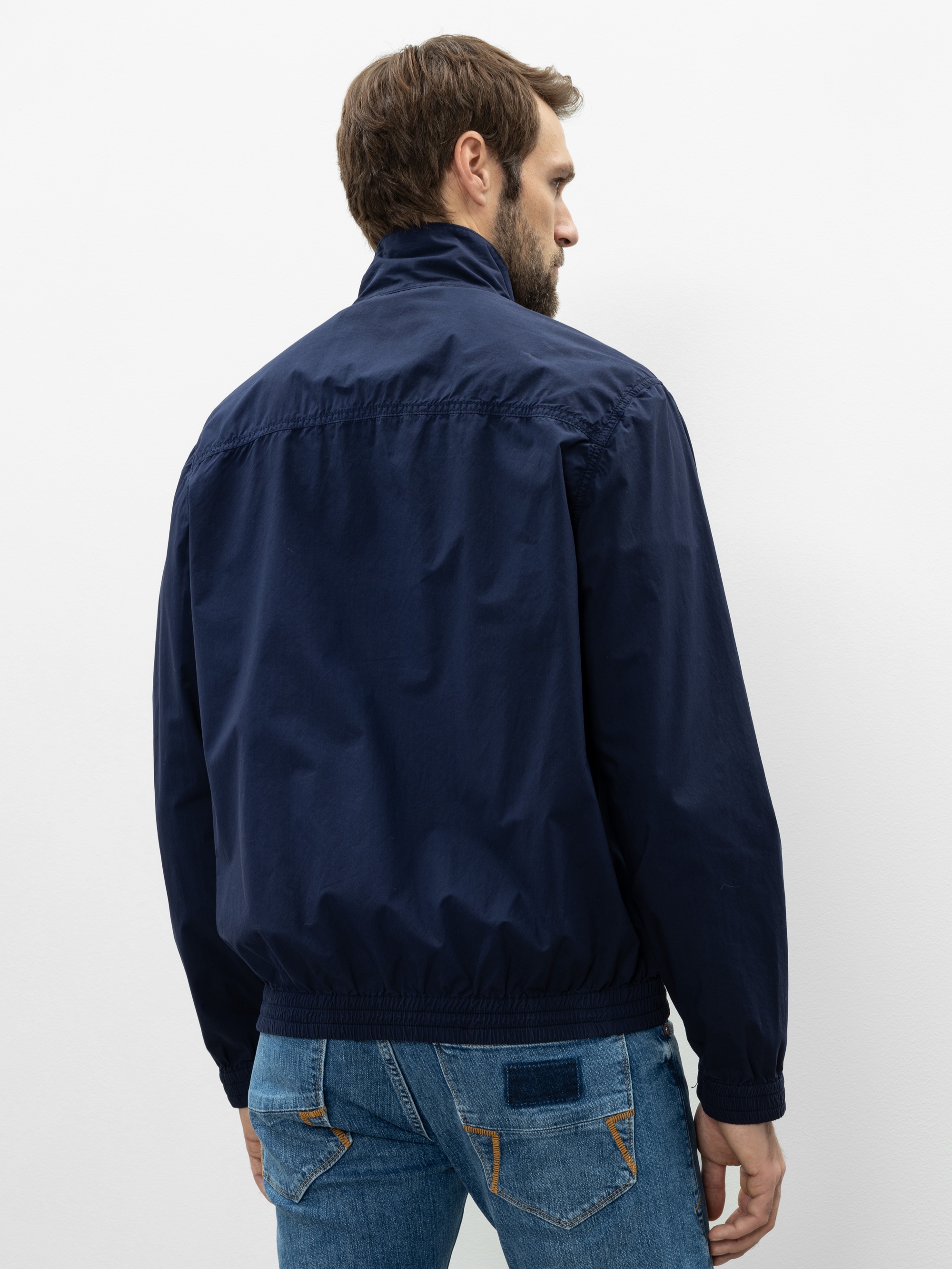 Ein Mann mit kurzen braunen Haaren steht mit dem Rücken zur Kamera, trägt eine marineblaue Jacke und blaue Jeans vor einem schlichten weißen Hintergrund.