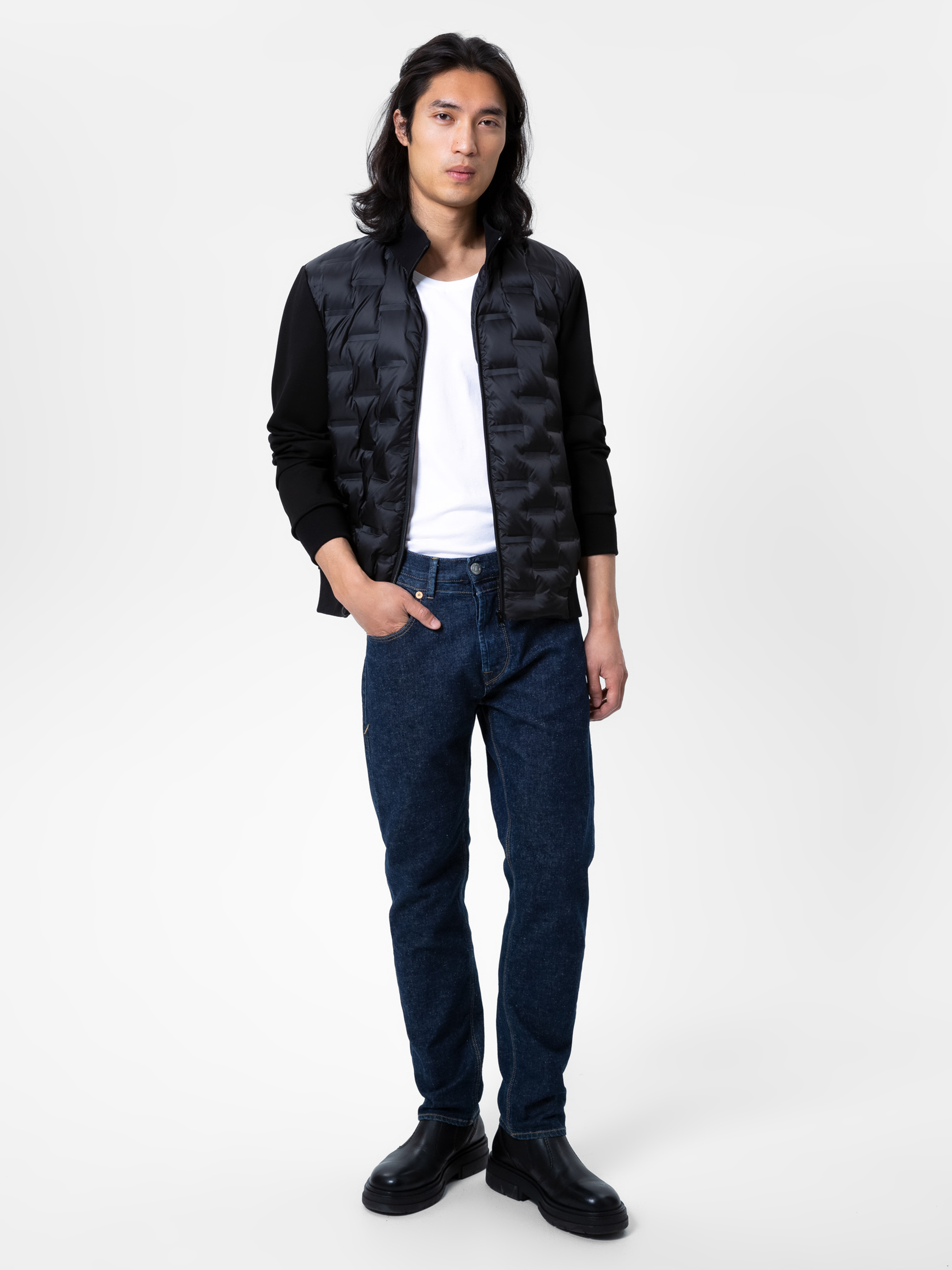 Eine Person mit langen dunklen Haaren trägt eine schwarze Jacke über einem weißen T-Shirt, blaue Jeans und schwarze Schuhe und steht vor einem schlichten weißen Hintergrund.