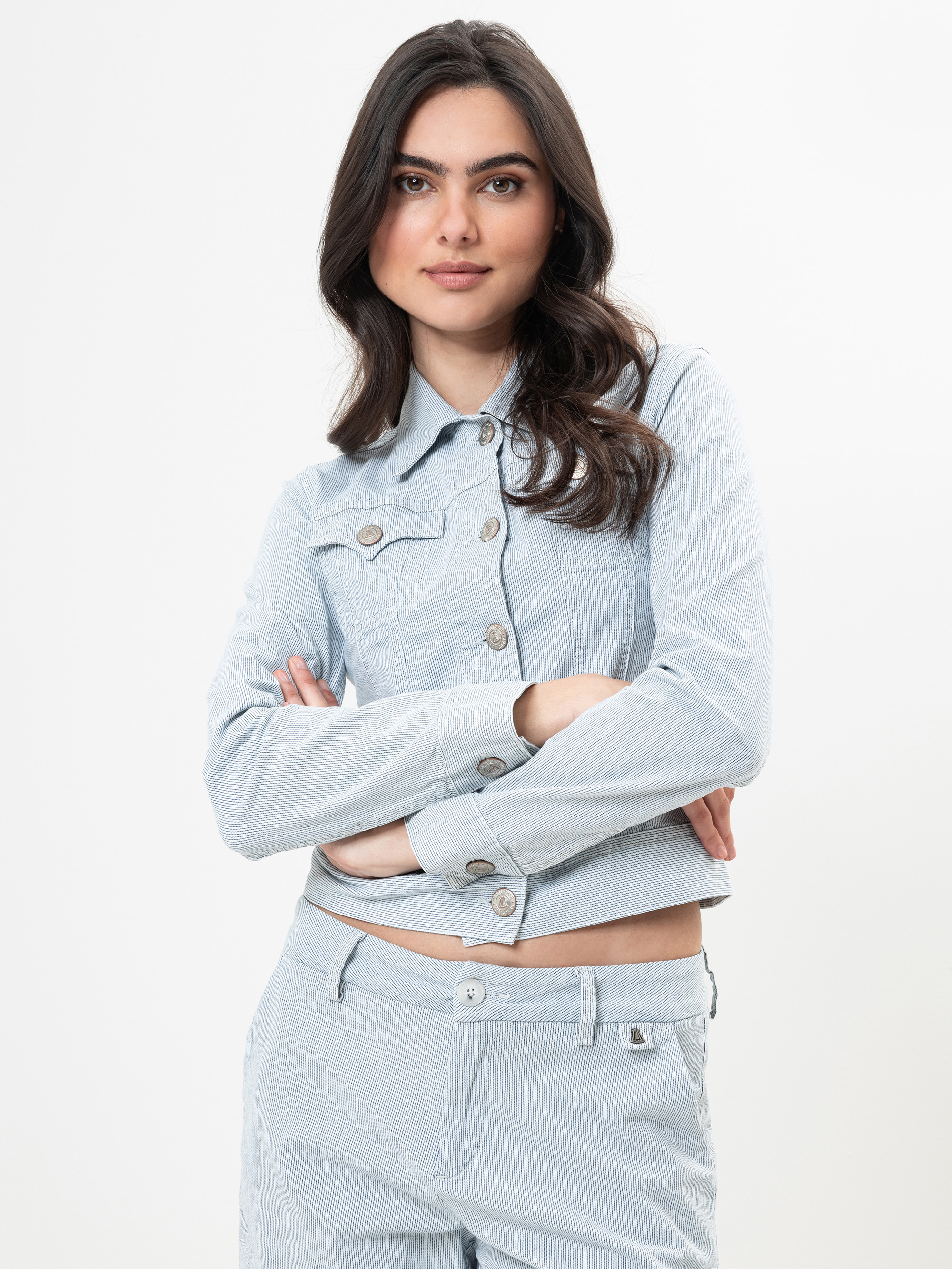 Eine Frau mit langen dunklen Haaren, die eine helle Jeansjacke und eine dazu passende Hose trägt, steht mit verschränkten Armen vor einem schlichten weißen Hintergrund und schaut selbstbewusst in die Kamera.