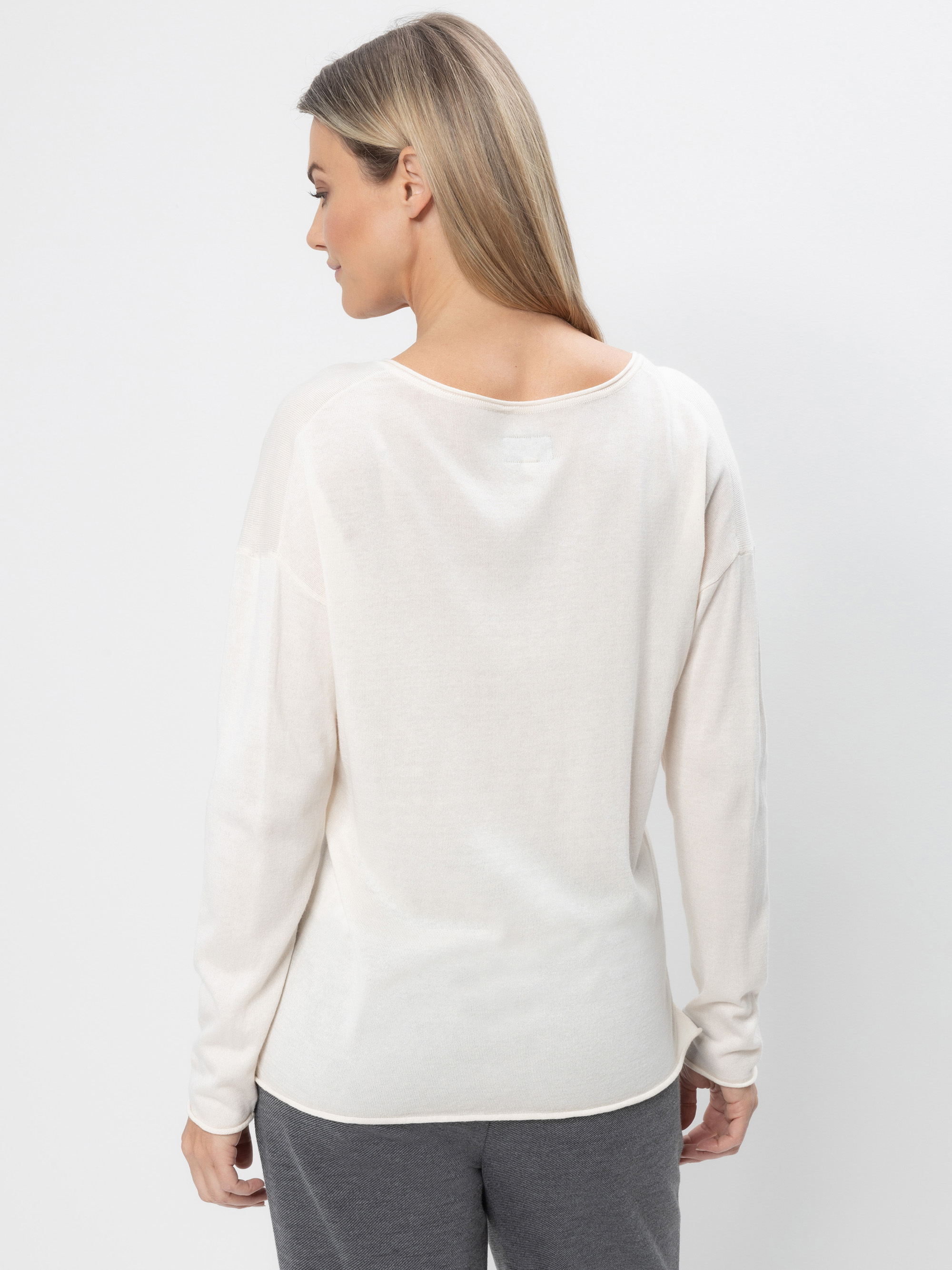 Eine Frau mit glattem, hellbraunem Haar trägt ein langärmeliges, cremefarbenes Oberteil und eine dunkelgraue Hose. Sie steht vor einem schlichten weißen Hintergrund und blickt von der Kamera weg.