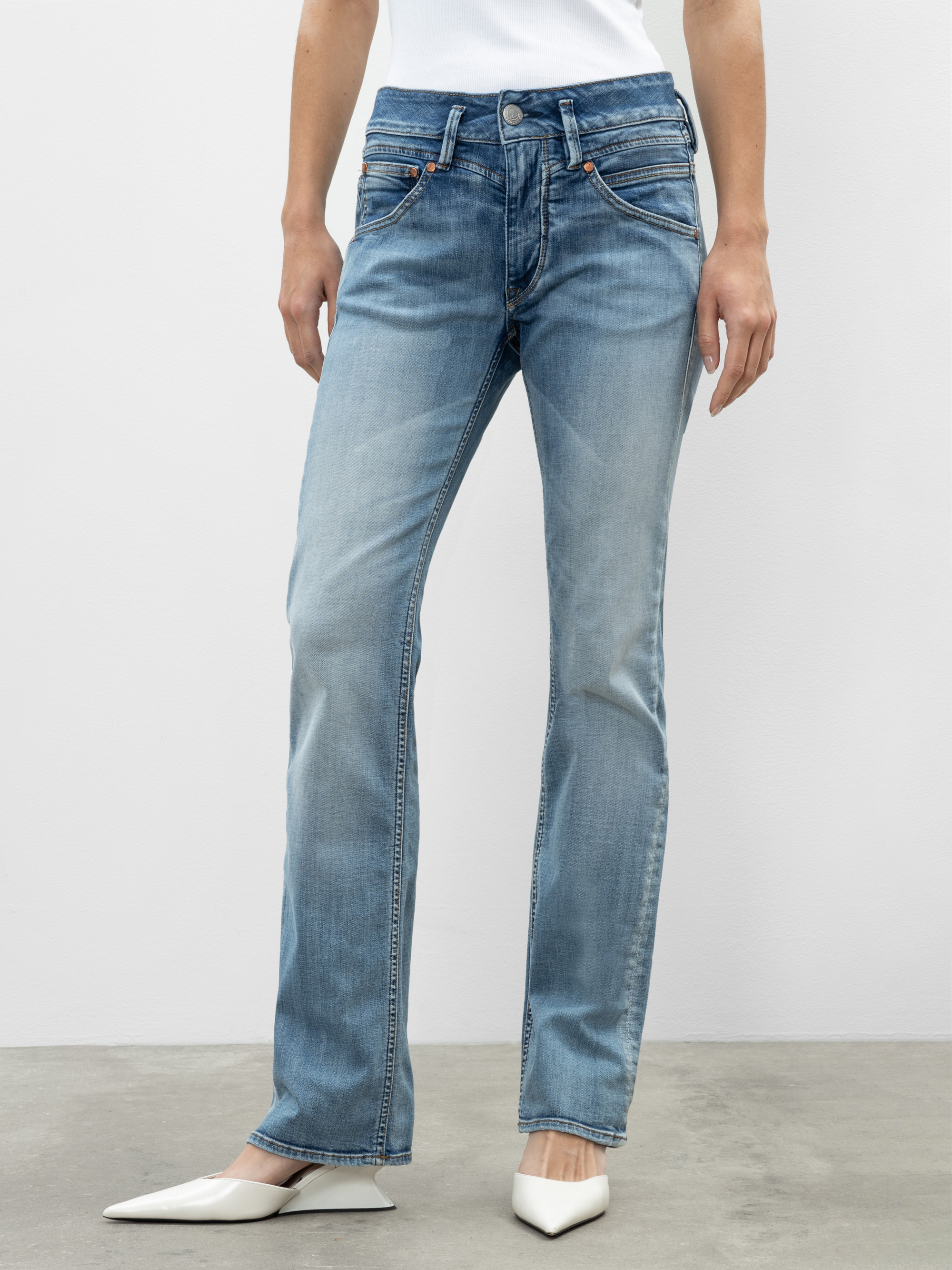Eine Person, die hellblaue Jeans mit geradem Bein, ein weißes Oberteil und weiße spitze Absätze trägt, steht auf einem grauen Boden vor einem einfarbig weißen Hintergrund.