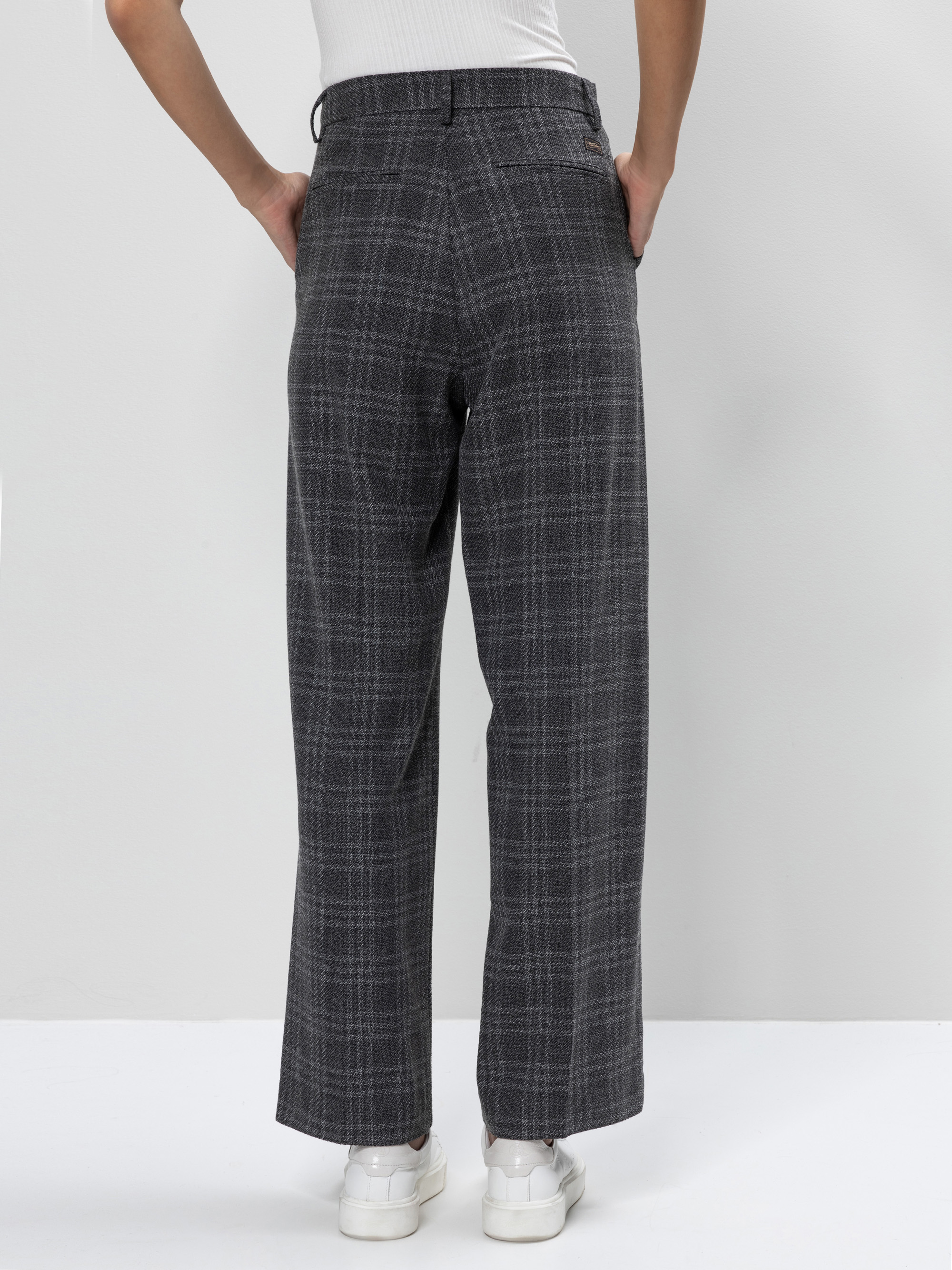 Eine Person steht mit dem Gesicht zur Wand, trägt eine grau karierte Hose mit weitem Bein, ein weißes ärmelloses Oberteil und weiße Turnschuhe, die Hände in den Taschen, vor einem schlichten hellen Hintergrund.