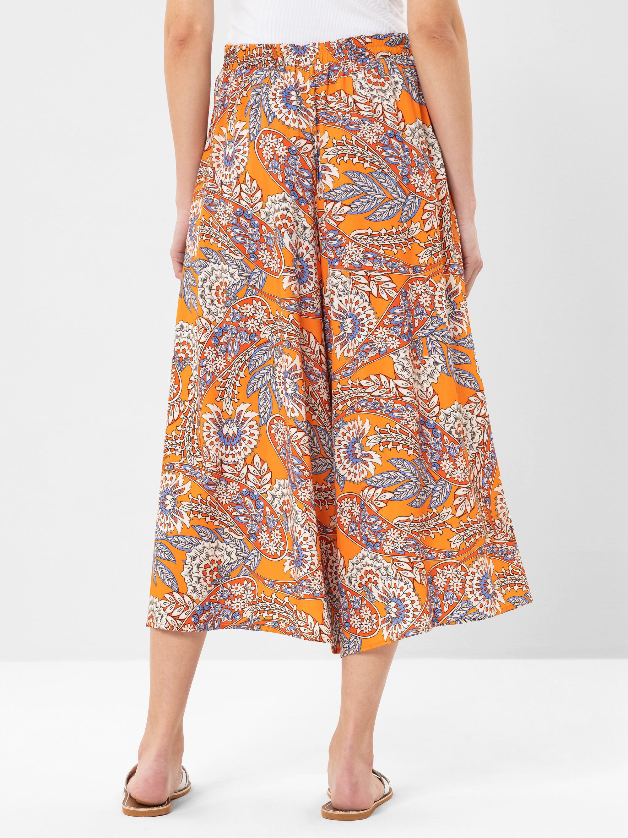 Eine Person steht mit dem Gesicht zur Wand und trägt eine orangefarbene Culotte-Hose mit buntem Blumen- und Blätterdruck, ein weißes Oberteil und beigefarbene flache Sandalen vor einem schlichten, hellen Hintergrund.