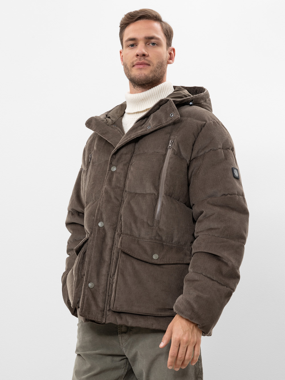 Ein Mann, der eine braune Cord-Pufferjacke mit großen Taschen und einer Kapuze trägt, steht vor einem schlichten weißen Hintergrund. Er trägt außerdem einen weißen Rollkragenpullover und eine olivgrüne Hose.