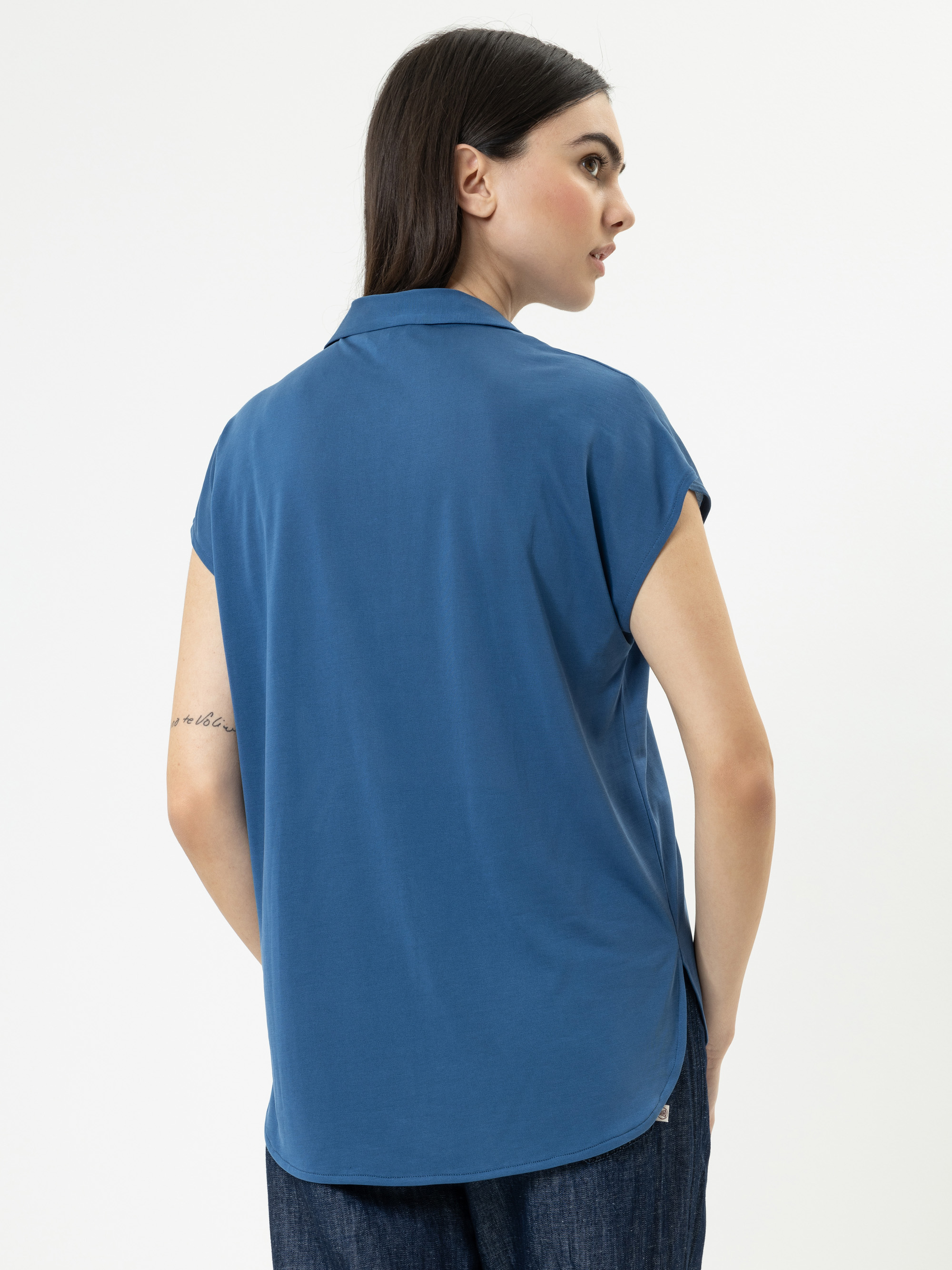 Eine Frau mit langen dunklen Haaren steht mit dem Gesicht zur Wand und trägt ein blaues kurzärmeliges Hemd mit Kragen und eine dunkle Hose. Ihr rechter Arm zeigt eine kleine Tätowierung. Der Hintergrund ist einfarbig weiß.