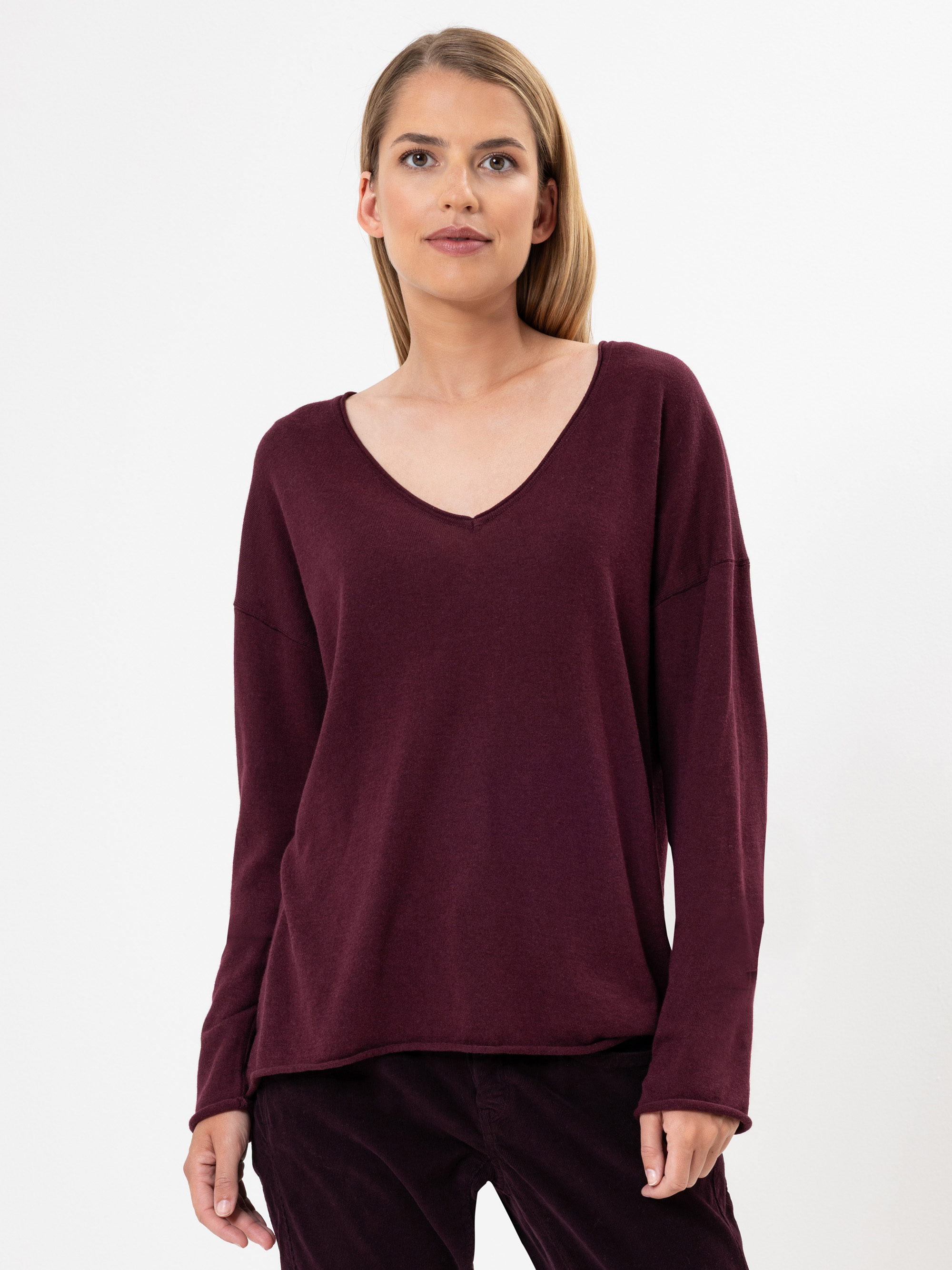 Eine Frau mit glattem blondem Haar trägt ein locker sitzendes, langärmeliges, dunkelbordeauxfarbenes Oberteil mit V-Ausschnitt und eine passende Hose. Sie steht vor einem schlichten weißen Hintergrund und blickt mit einem neutralen Gesichtsausdruck lei