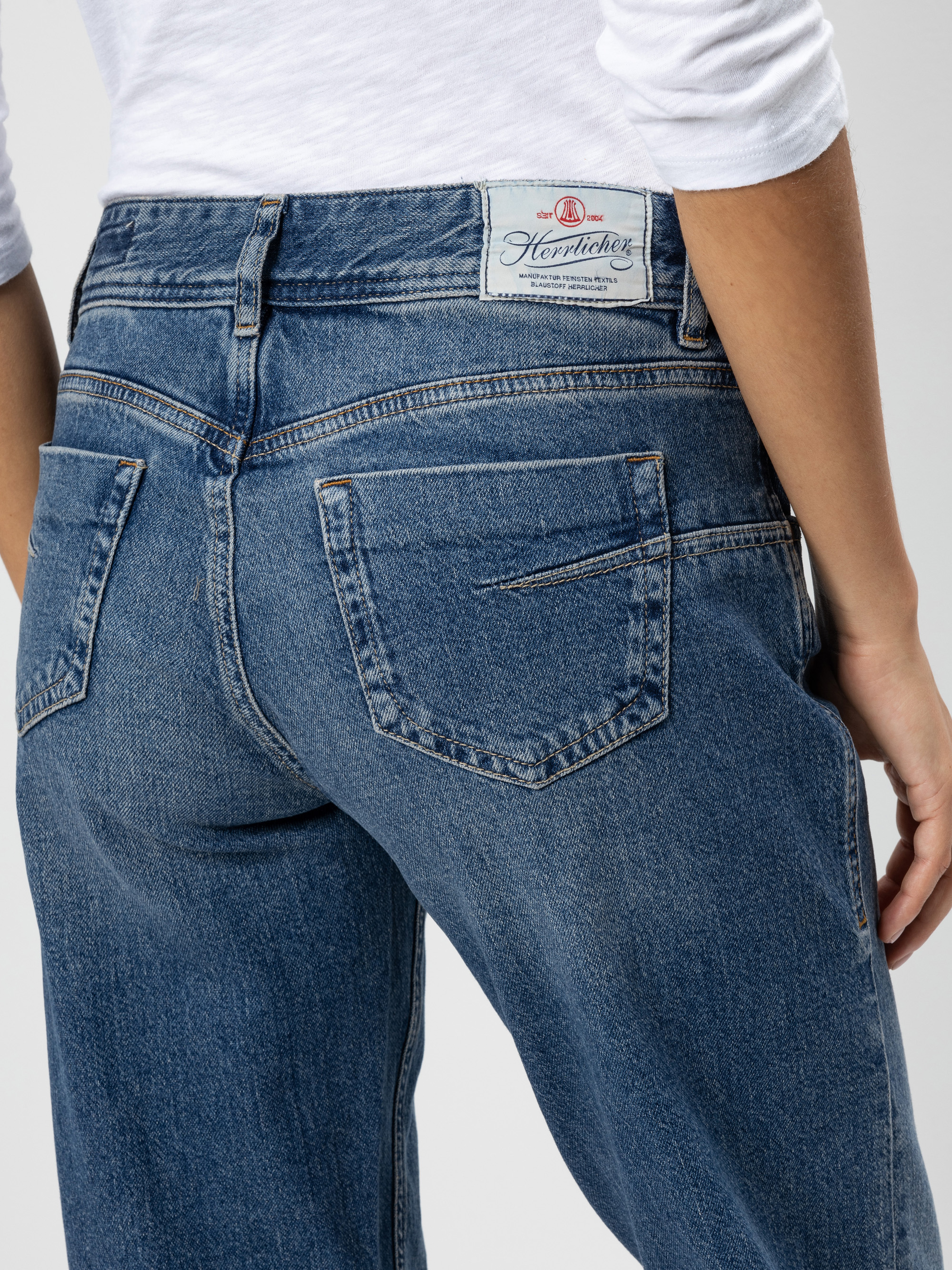 Person, die eine blaue Jeans mit hoher Taille und ein weißes Hemd trägt, von der Taille abwärts und von hinten gesehen. Die Jeans hat zwei Gesäßtaschen und ein Etikett am Bund.