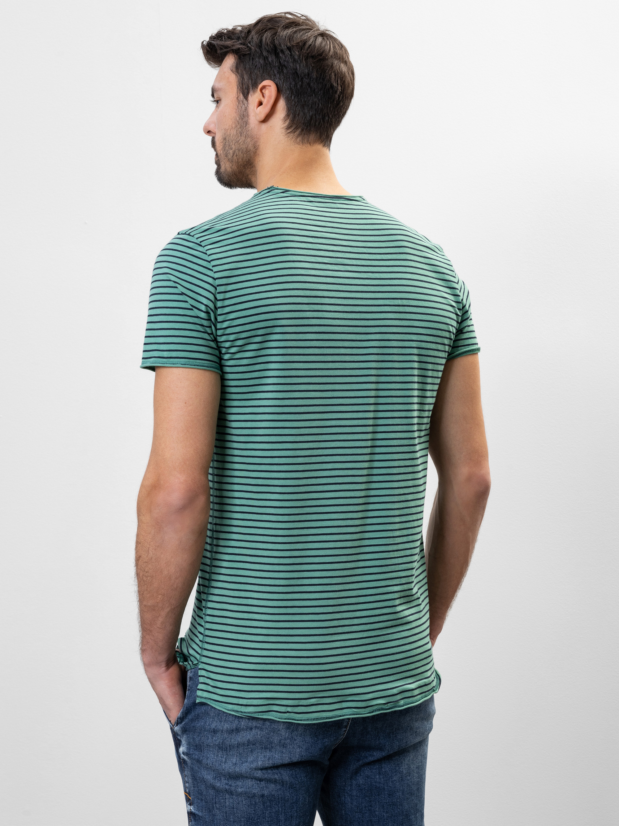 Ein Mann mit kurzen braunen Haaren steht mit dem Rücken zur Kamera, trägt ein grün-schwarz gestreiftes T-Shirt und blaue Jeans vor einem schlichten weißen Hintergrund.