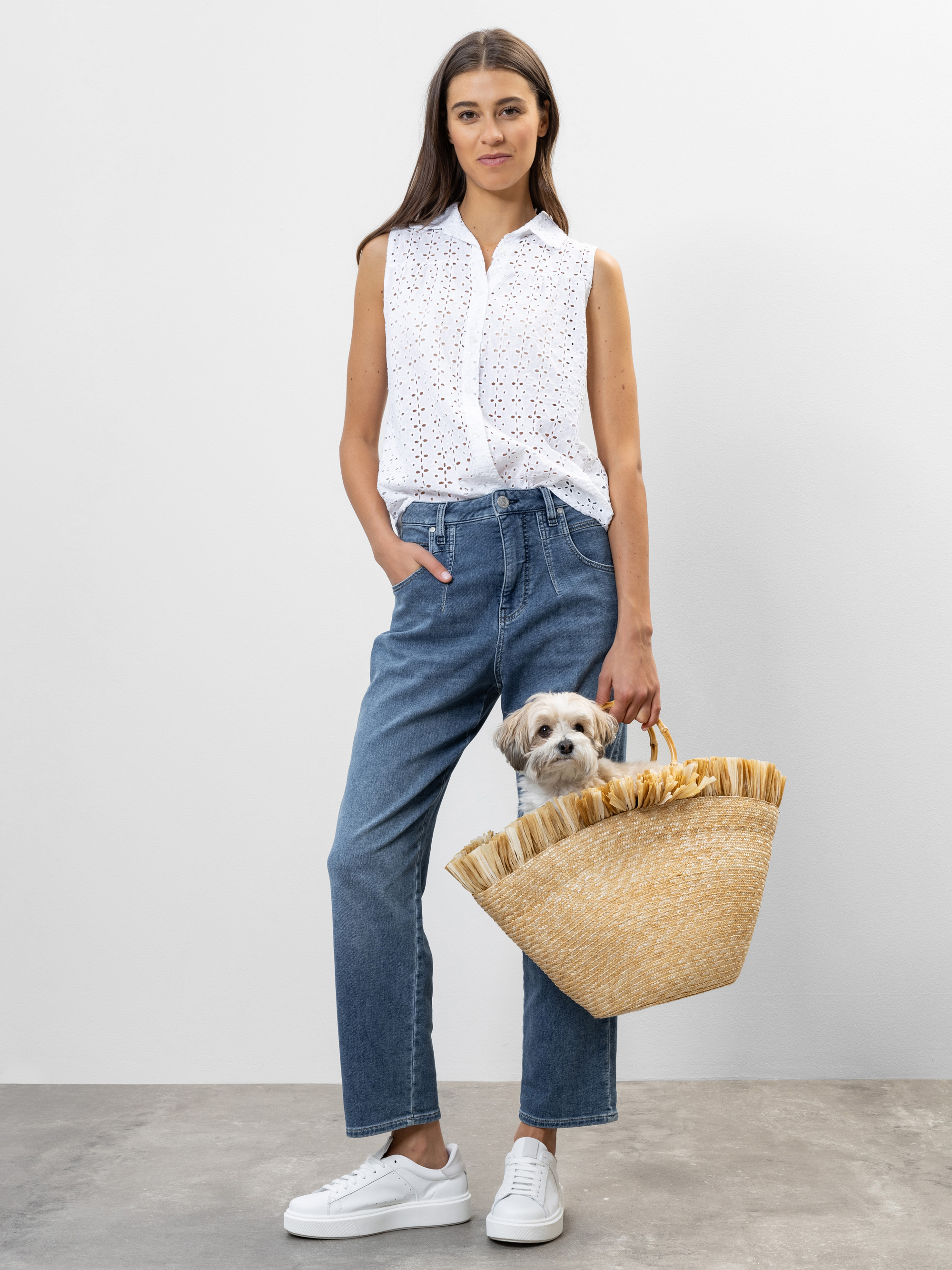 Eine Frau, die eine weiße ärmellose Bluse, blaue Jeans und weiße Turnschuhe trägt, steht vor einem schlichten Hintergrund und hält eine Tragetasche aus Stroh, in der ein kleiner Hund sitzt.