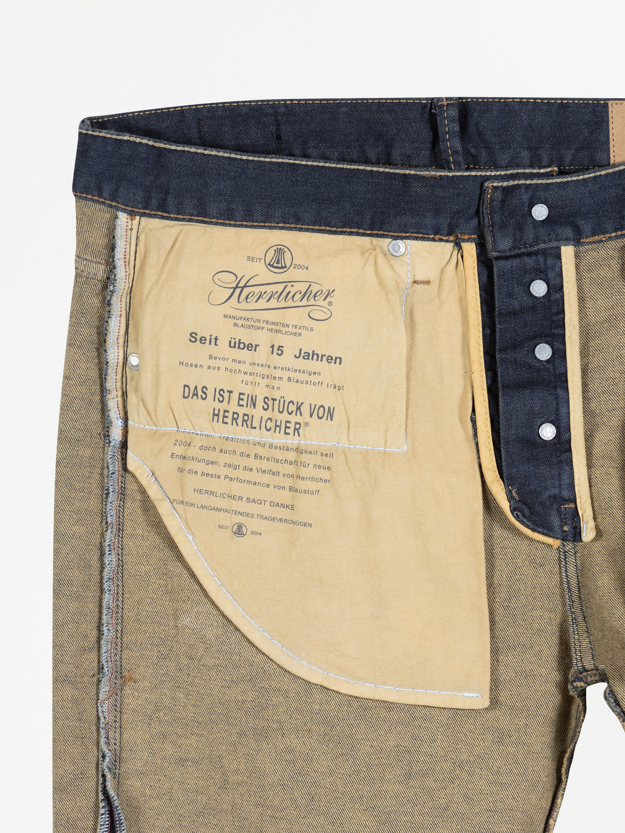 Nahaufnahme der Innenseite einer Jeans mit einem aufgedruckten Etikett in deutscher Sprache auf dem Taschenfutter, Markenzeichen Herrlicher, mit sichtbaren Nähten und Knöpfen am Bund und am Hosenschlitz.