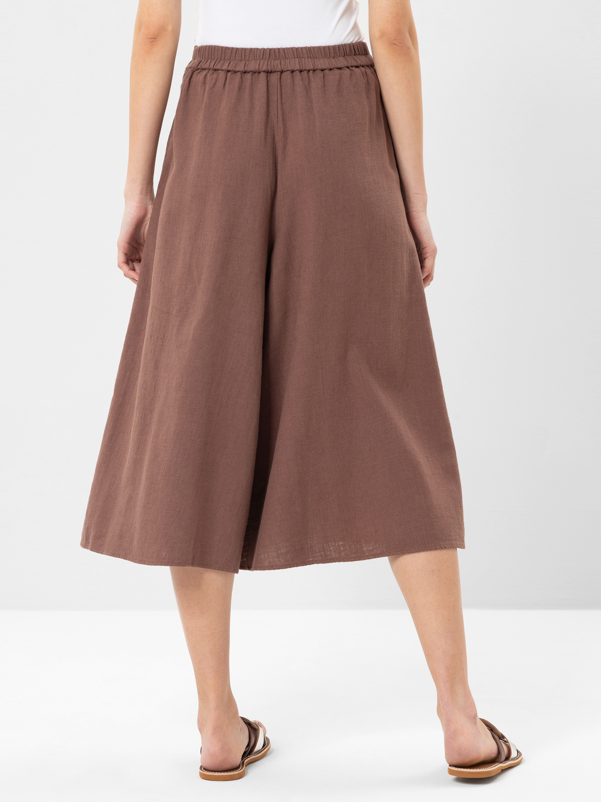 Eine Person steht mit weitem Bein, brauner Culotte-Hose und flachen Sandalen mit dem Gesicht nach unten. Die Hose hat einen elastischen Bund und die Person ist von der Taille abwärts vor einem einfarbigen Hintergrund abgebildet.