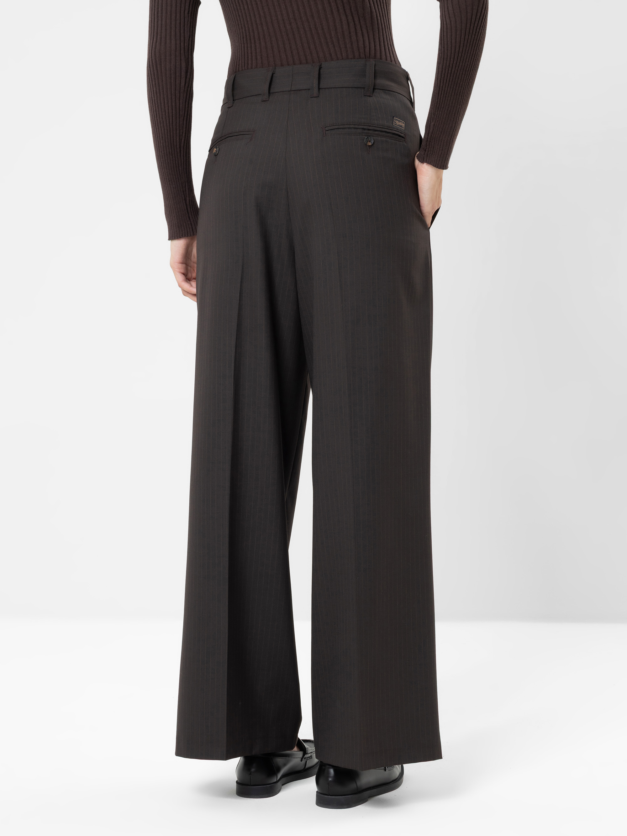 Eine Person, die mit dem Rücken zur Kamera steht und eine dunkle Hose mit hoher Taille und weitem Bein, ein dunkles, geripptes Oberteil, das in die Hose gesteckt ist, sowie schwarze Schuhe trägt, vor einem schlichten weißen Hintergrund. Die Hände lieg
