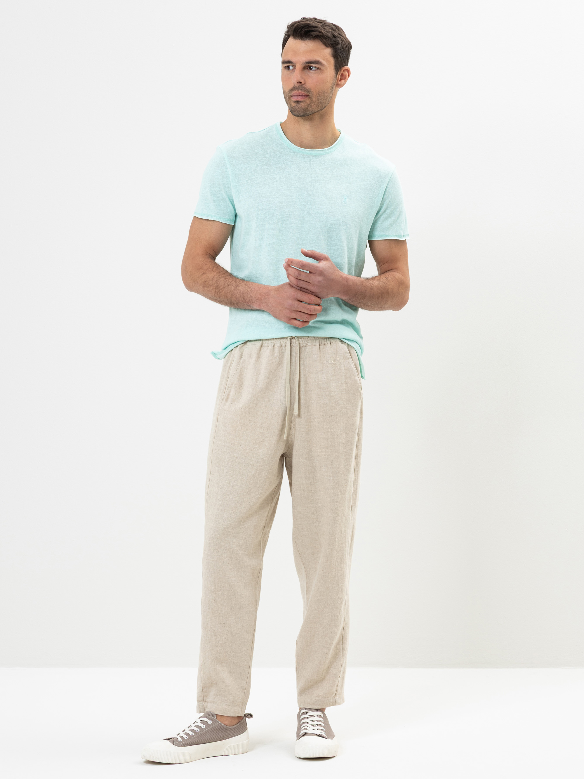 Ein Mann steht vor einem schlichten weißen Hintergrund und trägt ein helltürkisfarbenes T-Shirt, eine beigefarbene Hose mit Kordelzug und hellgraue Turnschuhe. Er blickt mit verschränkten Händen zur Seite.