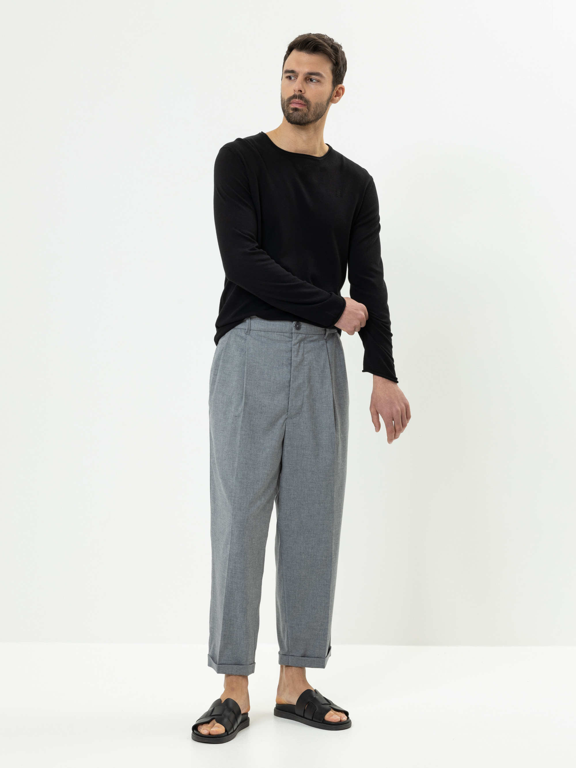 Ein Mann steht vor einem schlichten weißen Hintergrund und trägt ein schwarzes langärmeliges Hemd, eine hellgraue Bundfaltenhose mit Bündchen und schwarze Sandalen. Er blickt leicht zur Seite und hat einen Arm über den Körper gebogen.