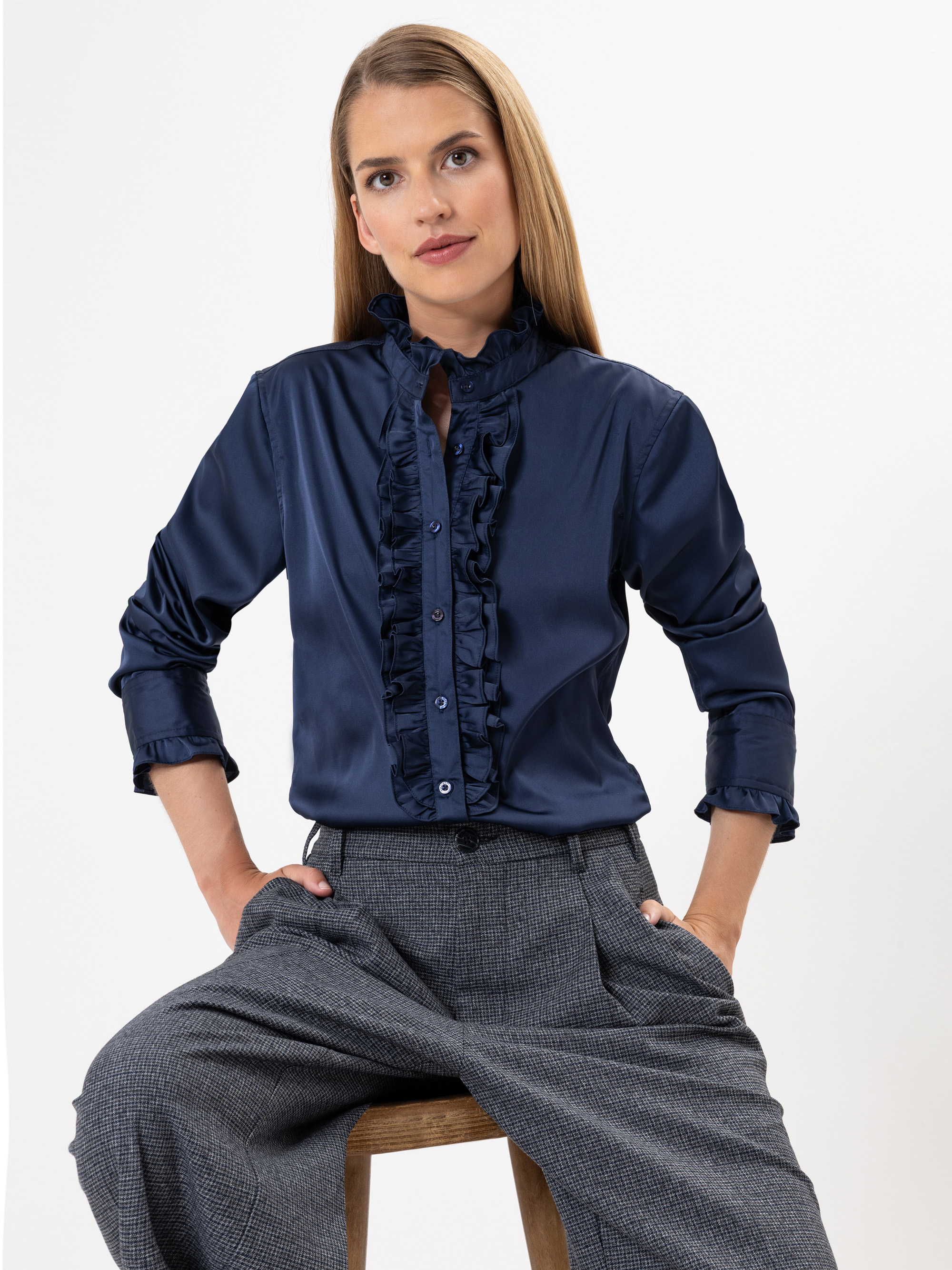 Eine Frau mit glattem, hellbraunem Haar sitzt auf einem Hocker und trägt eine marineblaue Rüschenbluse mit langen Ärmeln und eine graue Hose mit hoher Taille. Sie hat einen neutralen Gesichtsausdruck und einen weißen Hintergrund.