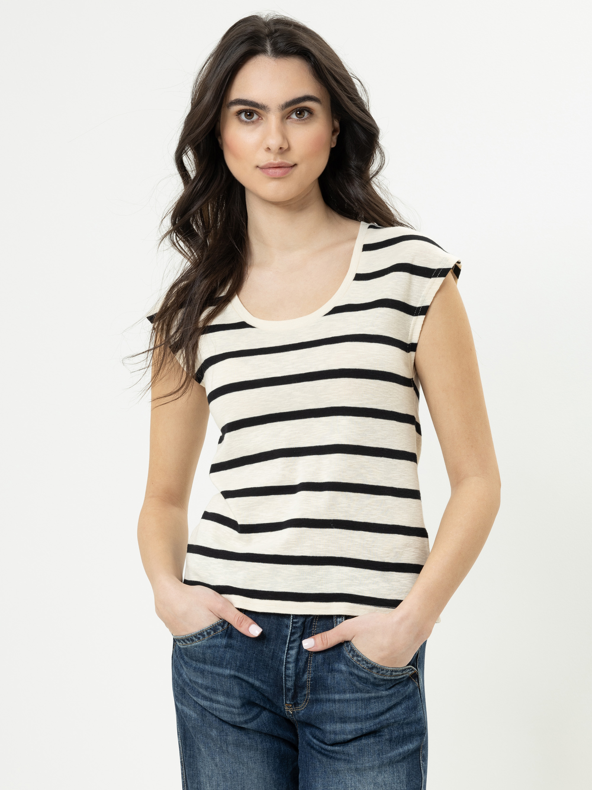 Eine Frau mit langen braunen Haaren trägt ein cremefarbenes, kurzärmeliges Oberteil mit schwarzen Querstreifen und blaue Jeans. Sie steht mit den Händen in den Taschen vor einem schlichten weißen Hintergrund.