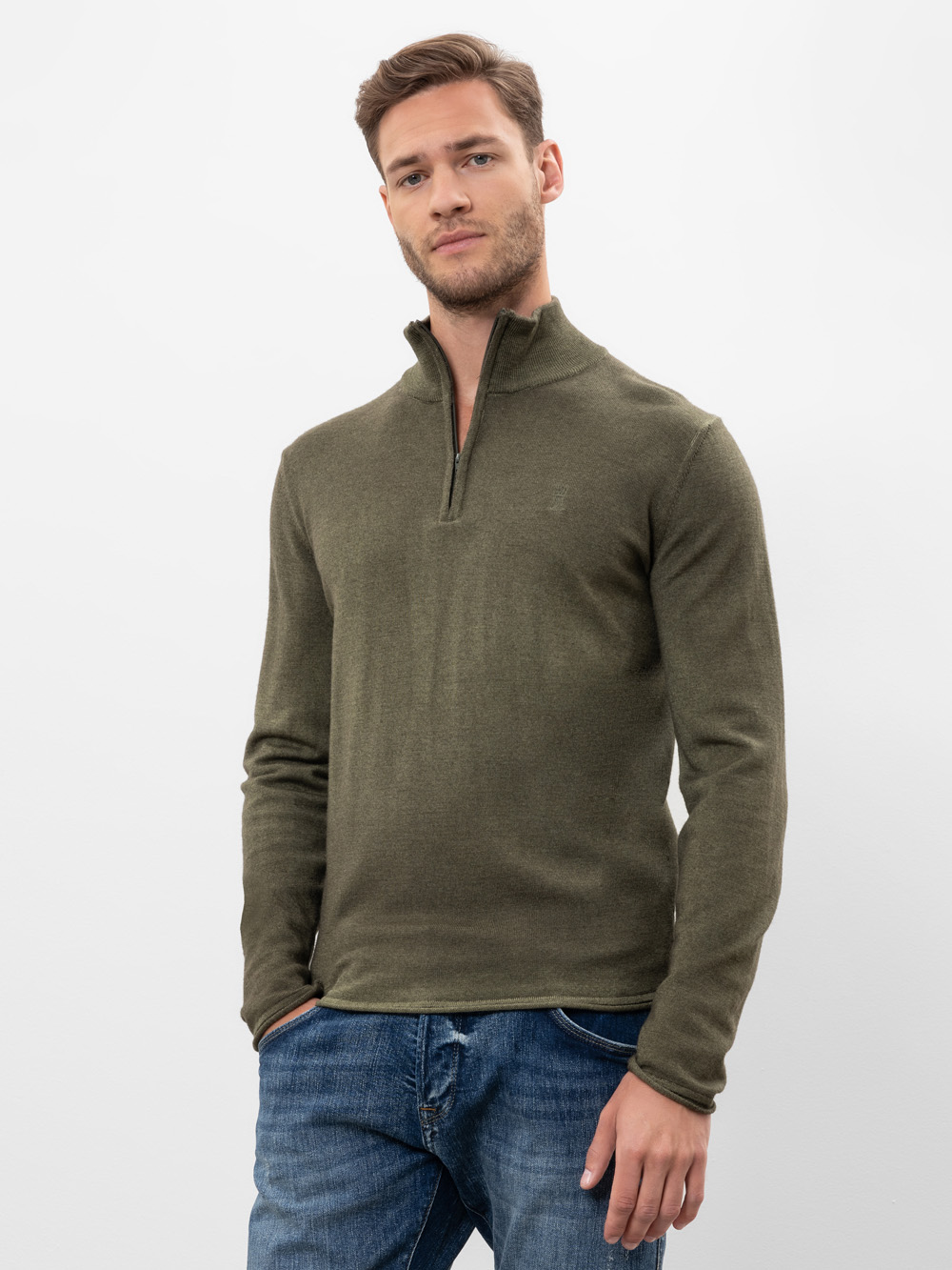 Ein Mann, der einen khakigrünen Pullover mit vierteljährlichem Reißverschluss und blaue Jeans trägt, steht vor einem schlichten weißen Hintergrund und wirkt entspannt.