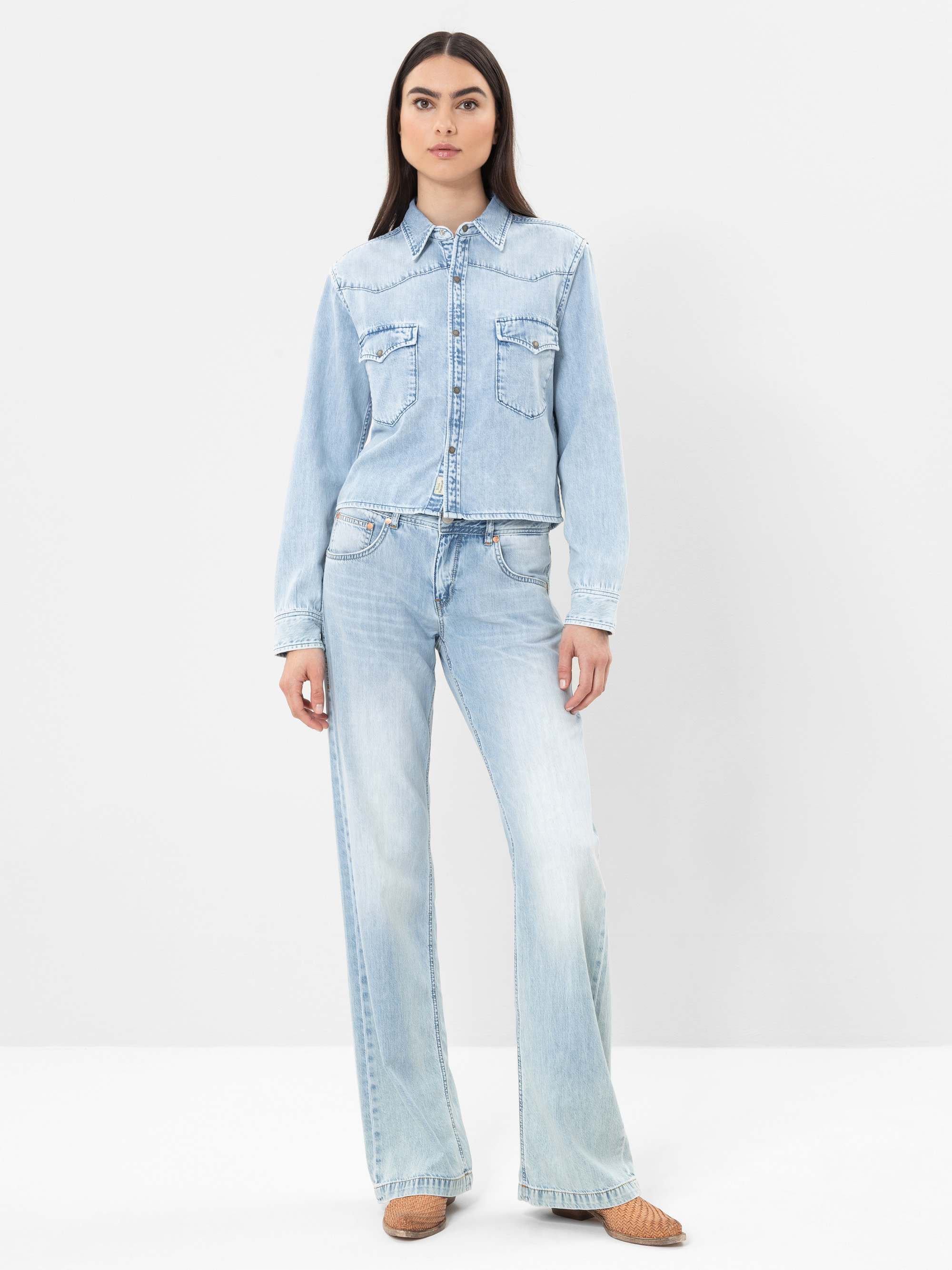 Eine Frau steht vor einem schlichten weißen Hintergrund und trägt ein hellblaues Jeanshemd mit Vordertaschen und dazu passende hellblaue Jeans mit weitem Bein. Sie trägt außerdem hellbraune gewebte Schuhe.