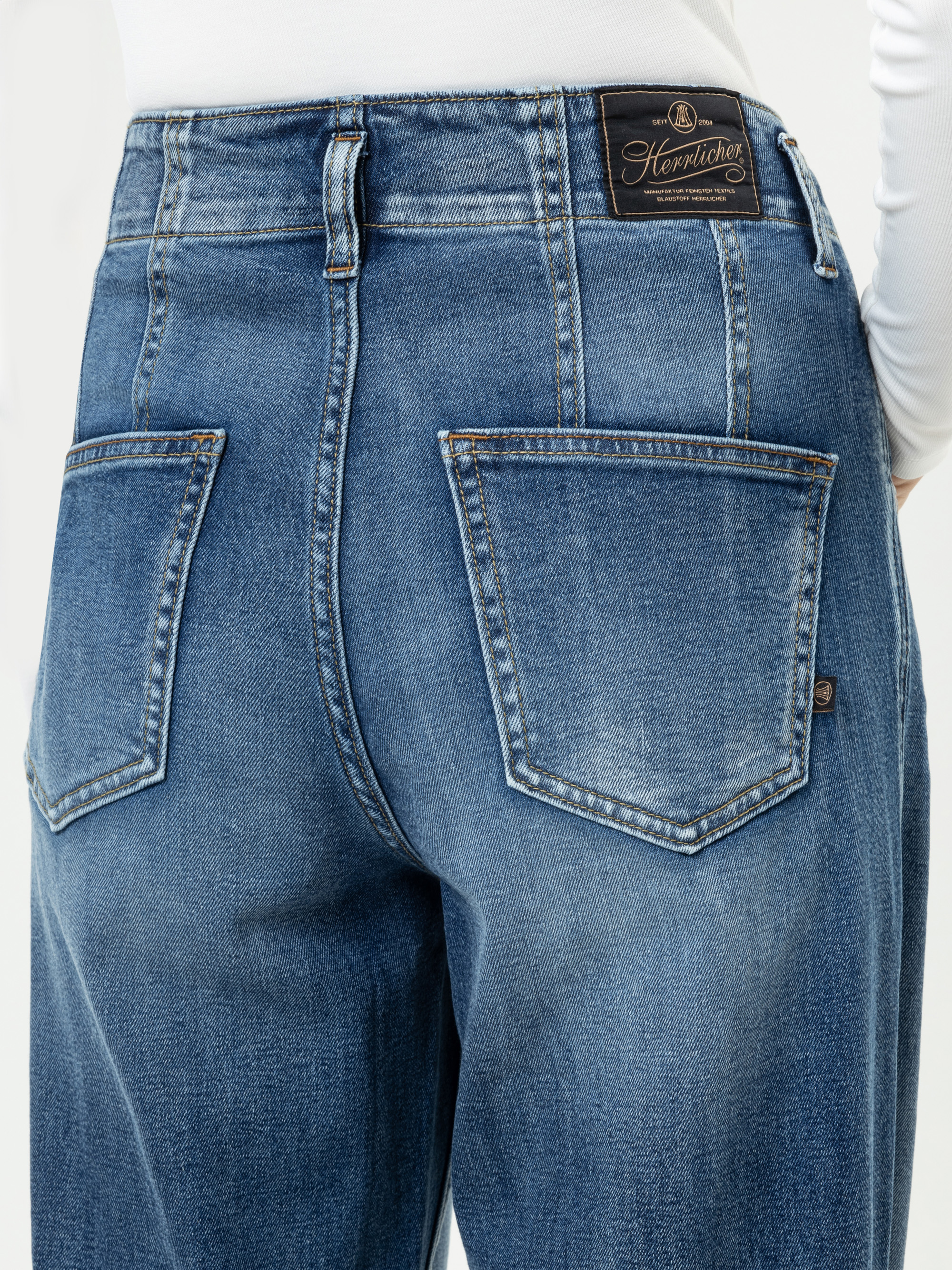 Nahaufnahme einer Person, die eine blaue Denim-Jeans und ein weißes Oberteil trägt. Zu sehen ist die Rückseite der Jeans mit zwei Gesäßtaschen und einem schwarzen Henrlicher-Markenaufnäher am Bund.
