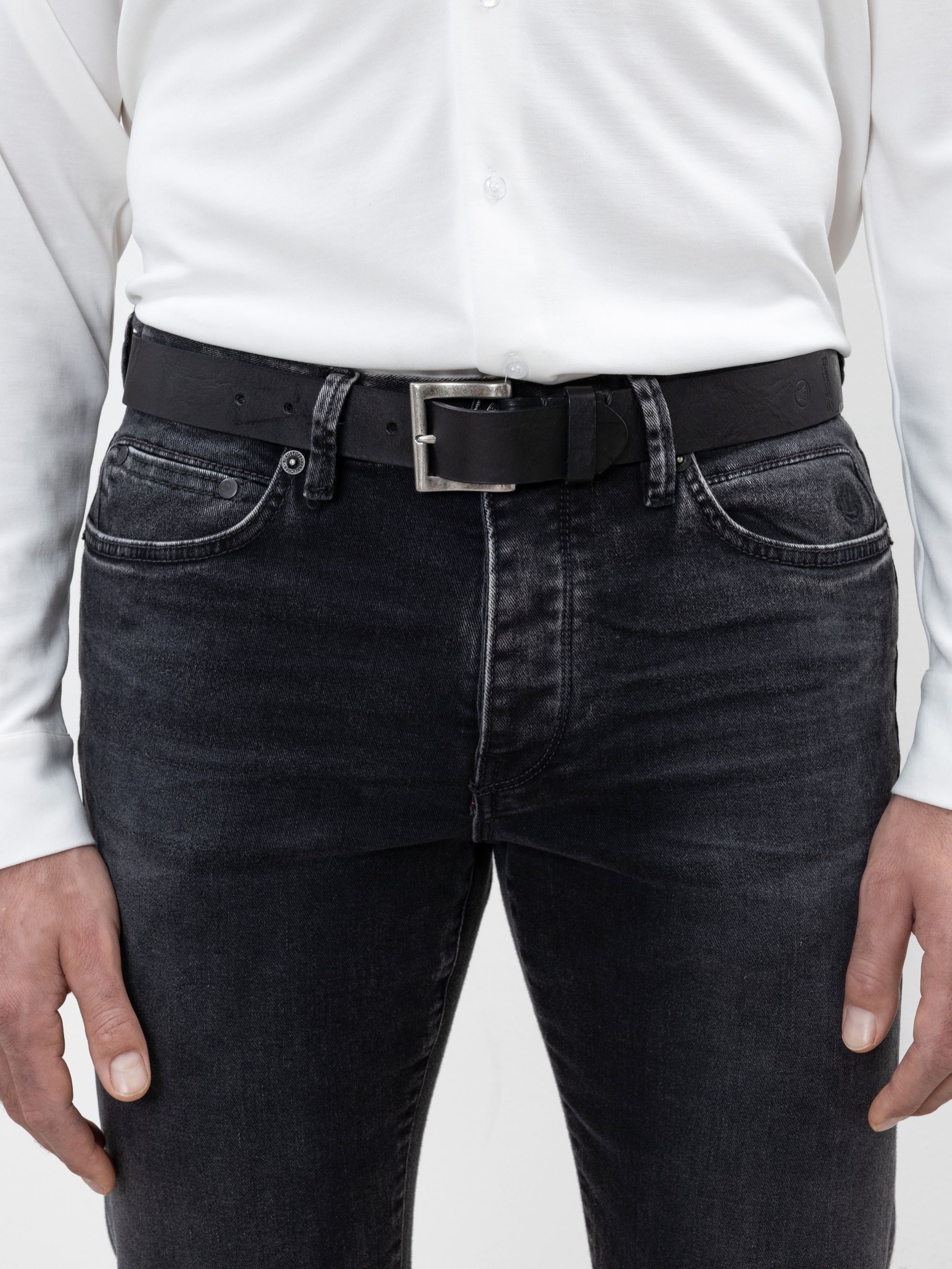 Eine Person, die ein weißes Button-up-Hemd, einen schwarzen Ledergürtel und eine dunkle Jeans trägt, die von der Taille bis zur Mitte der Oberschenkel reicht und deren Hände locker an der Seite liegen.