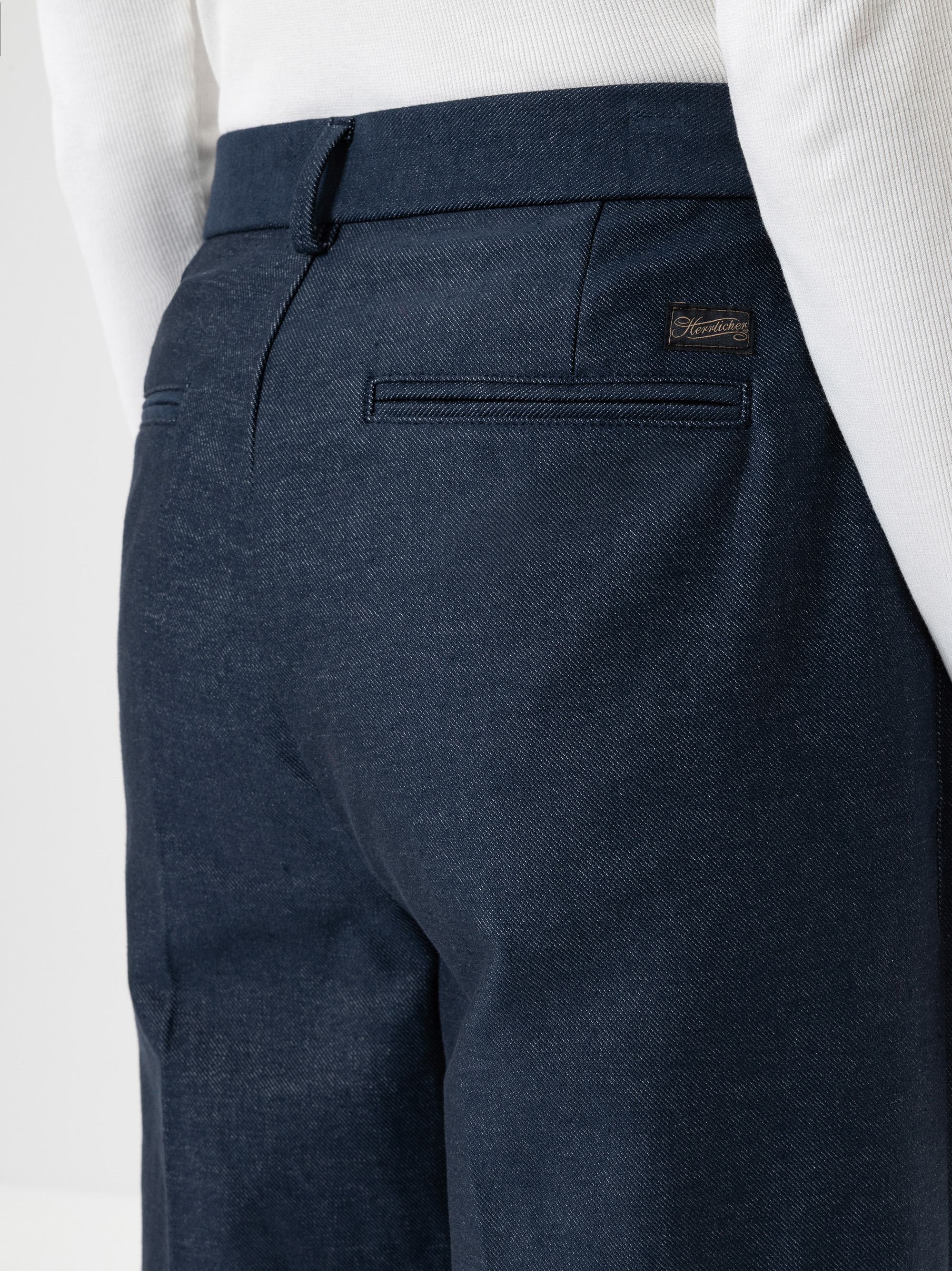 Nahaufnahme einer Person, die eine dunkelblaue maßgeschneiderte Hose und ein weißes Hemd trägt, wobei der Bereich der Gesäßtasche mit einem kleinen Markenetikett über der Tasche zu sehen ist.