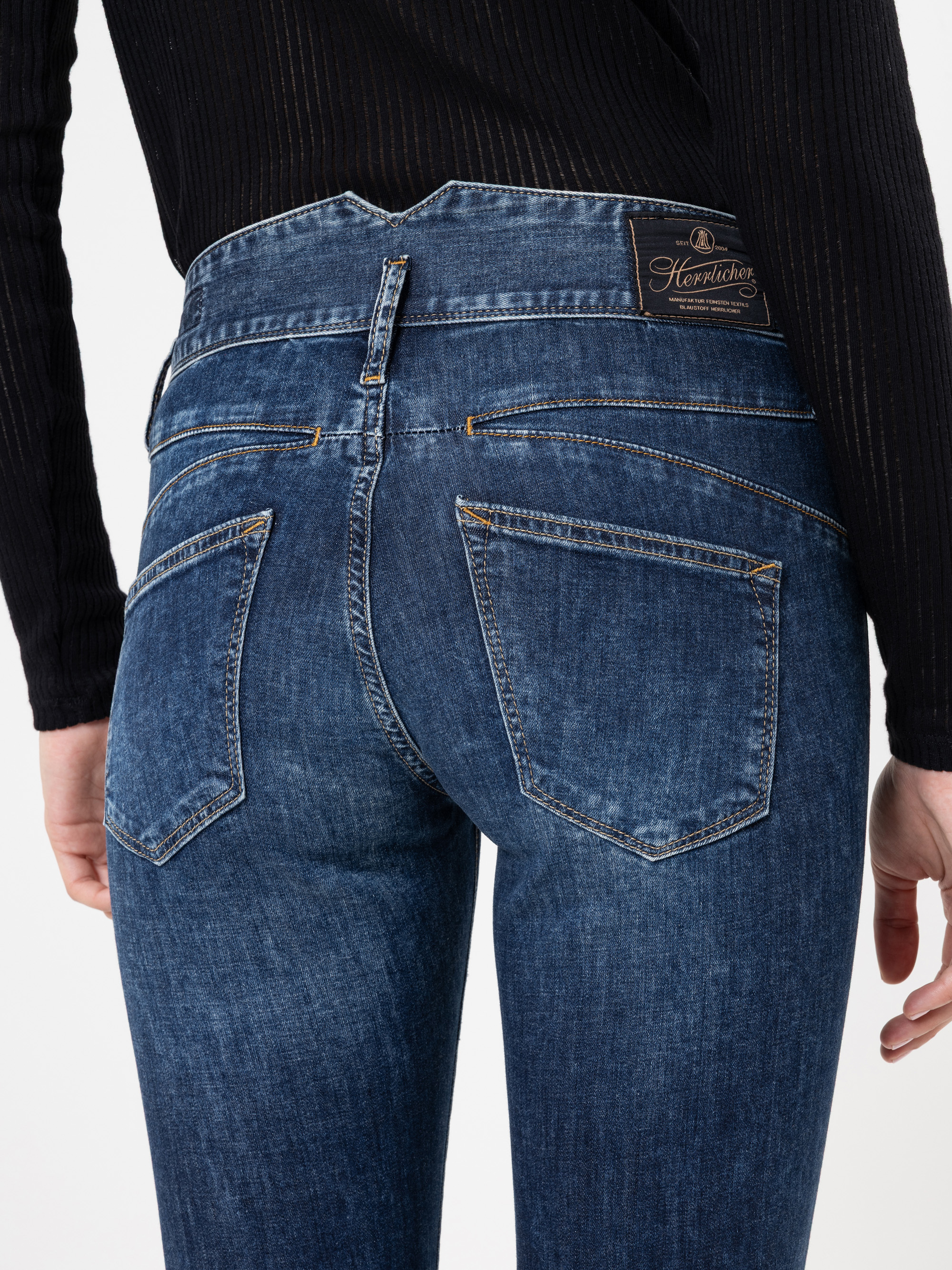 Eine Person trägt eine blaue Jeans mit hoher Taille und ein schwarzes langärmeliges Rippenoberteil, von hinten gesehen. Die Jeans hat ein aufgenähtes Etikett am Bund und sichtbare Taschen.