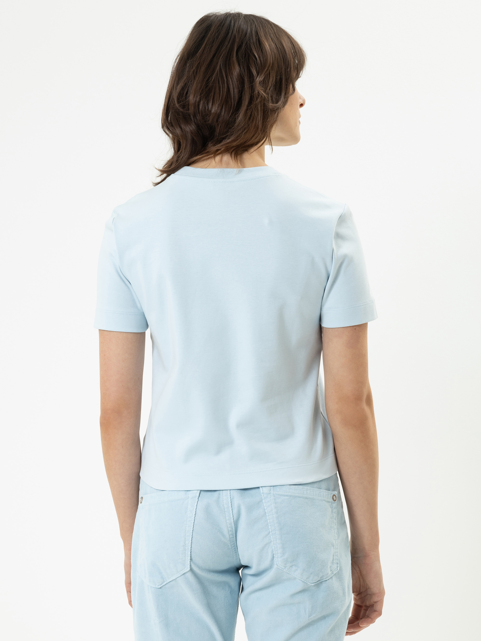 Eine Frau mit schulterlangen braunen Haaren steht mit dem Rücken zur Kamera, trägt ein hellblaues kurzärmeliges Hemd und hellblaue Jeans vor einem schlichten weißen Hintergrund.