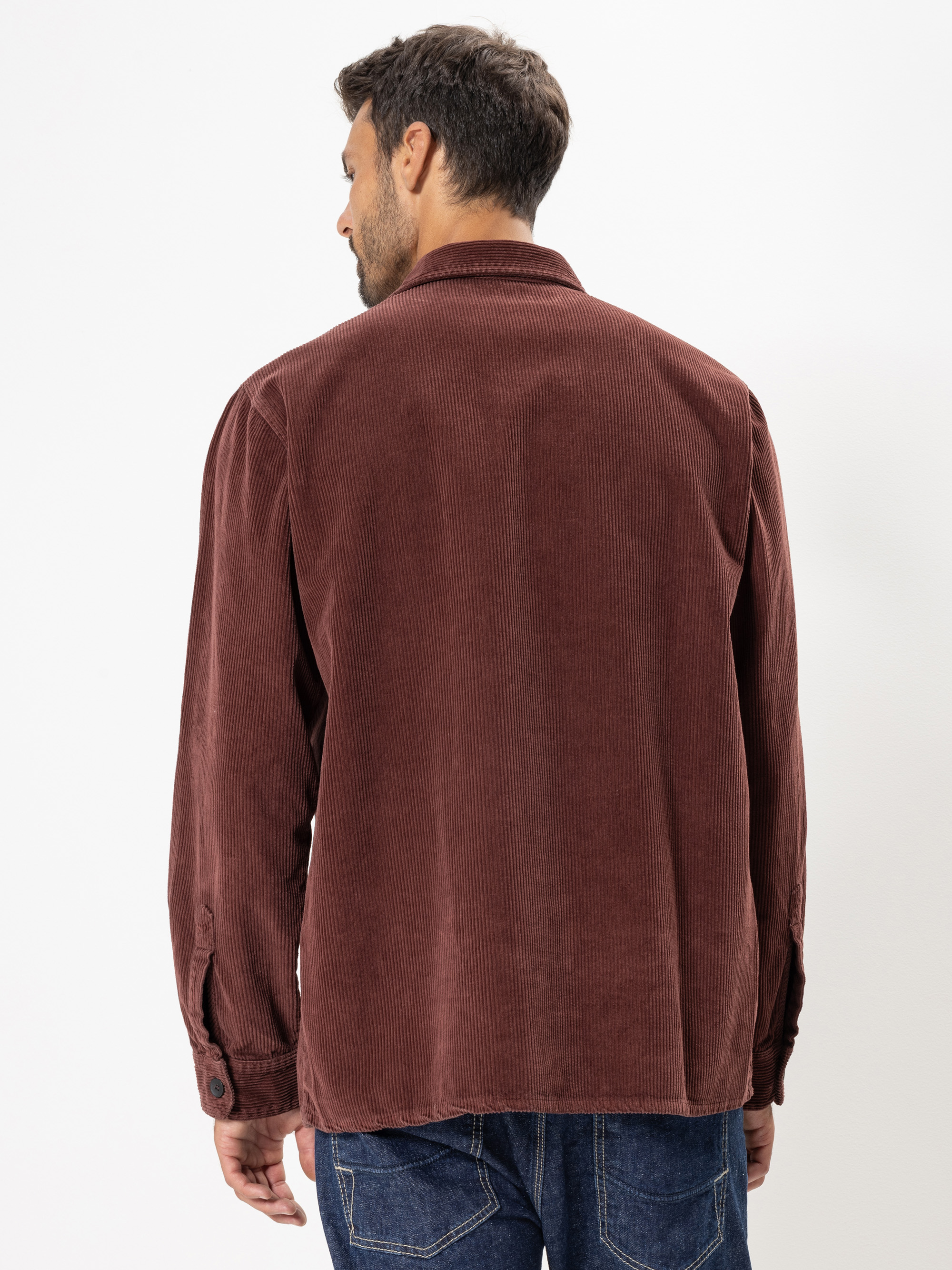 Ein Mann wird von hinten gezeigt, der ein langärmeliges, braunes Cordhemd und blaue Jeans trägt und vor einem schlichten weißen Hintergrund steht.
