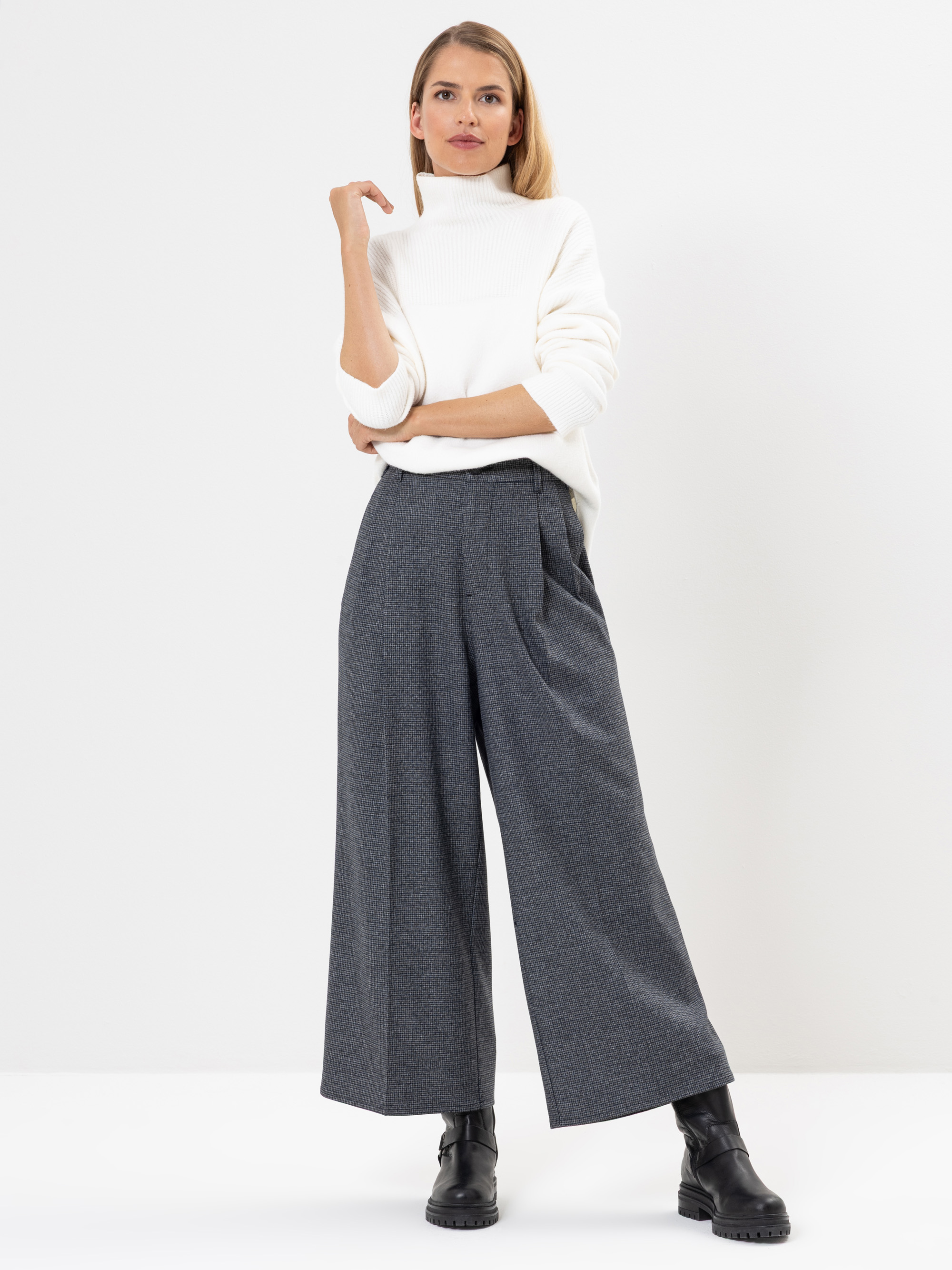 Eine Frau steht vor einem schlichten weißen Hintergrund und trägt einen weißen Rollkragenpullover, eine graue Hose mit weitem Bein und schwarze Stiefel. Sie hat einen Arm angewinkelt, die Hand am Kinn und blickt selbstbewusst nach vorne.