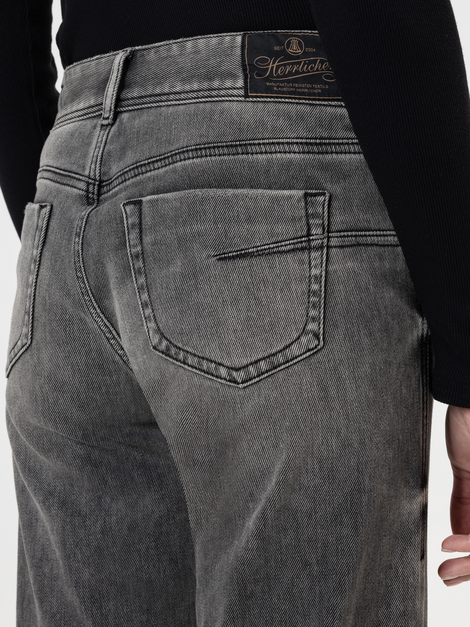 Nahaufnahme einer Person, die eine verblichene graue Jeans mit sichtbaren Nähten und Gesäßtaschen trägt. Die Jeans hat einen braunen Lederaufnäher am Bund mit Herrlicher-Schriftzug. Die Person trägt außerdem ein schwarzes Oberteil.