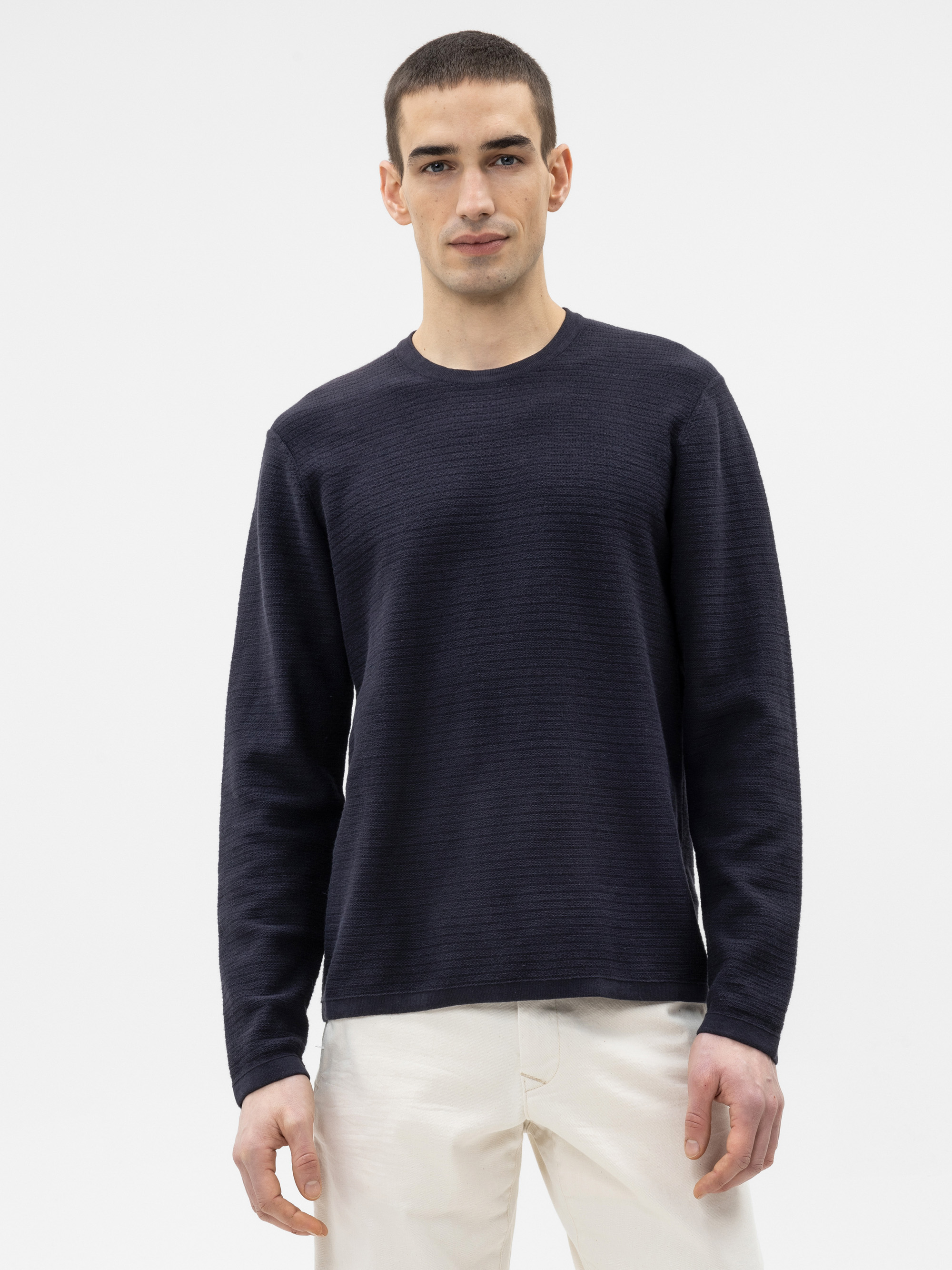 Ein Mann mit kurzen dunklen Haaren, der ein strukturiertes, langärmeliges marineblaues Hemd und eine weiße Hose trägt, steht vor einem schlichten weißen Hintergrund und blickt mit neutraler Miene nach vorn.