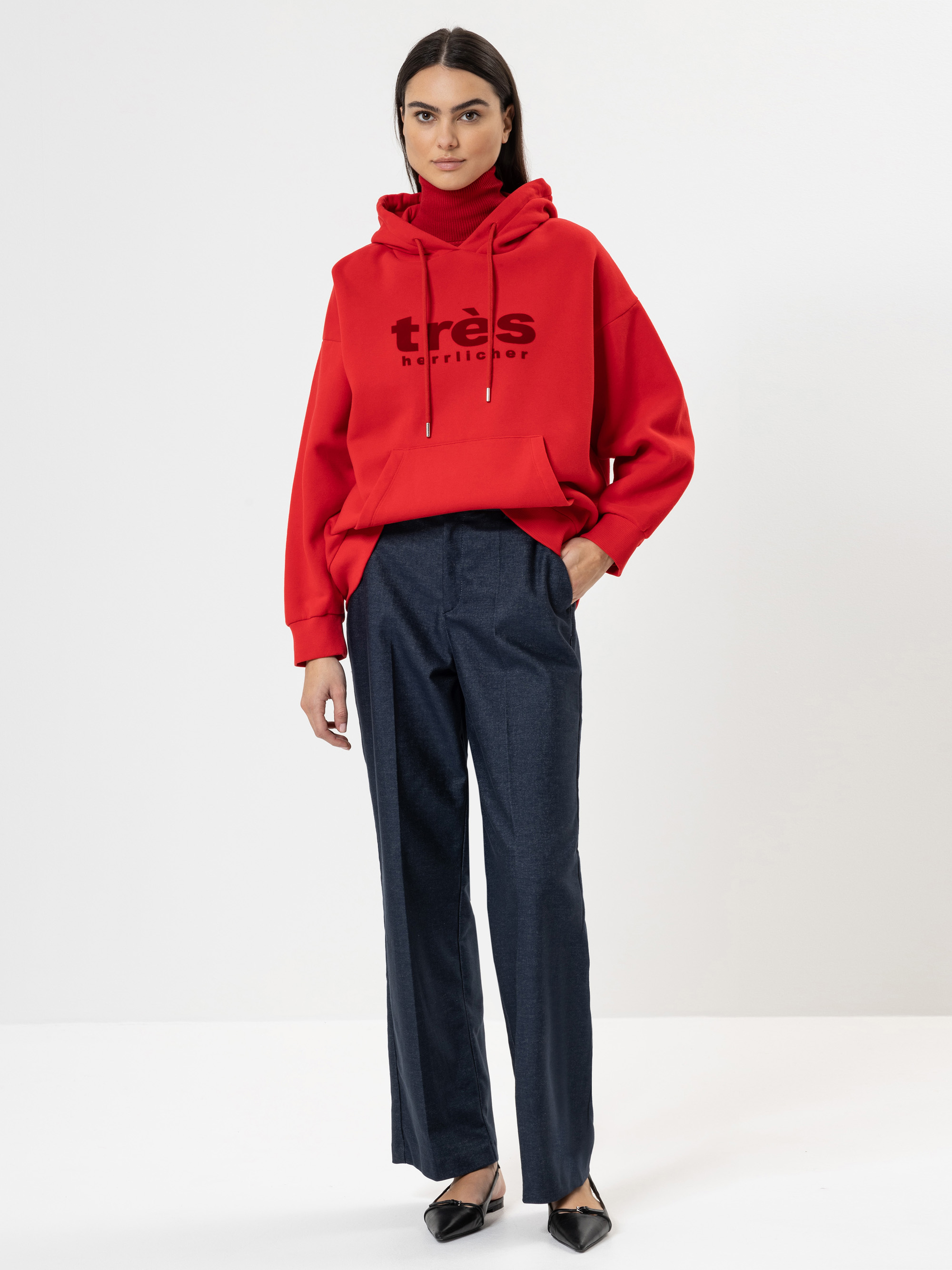 Eine Frau steht vor einem schlichten weißen Hintergrund und trägt einen roten Kapuzenpulli mit dem Aufdruck très harris, eine dunkelblaue Anzughose und schwarze, spitz zulaufende Schuhe. Sie hat glattes dunkles Haar und einen neutralen Gesichtsausdruck
