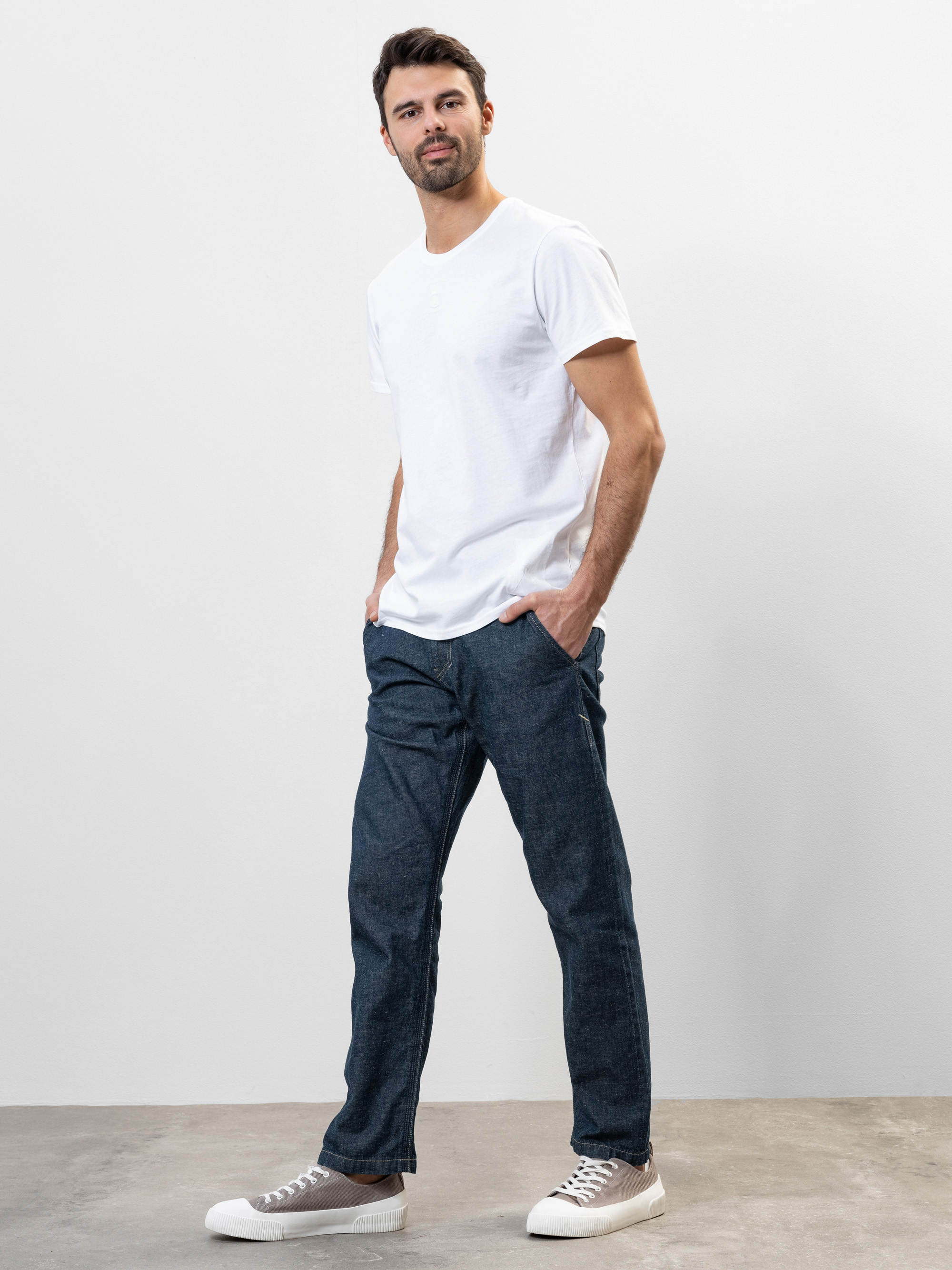 Ein Mann steht lässig vor einem schlichten weißen Hintergrund, trägt ein weißes T-Shirt, blaue Jeans und weiße Turnschuhe, die Hände in den Taschen und einen entspannten Gesichtsausdruck.