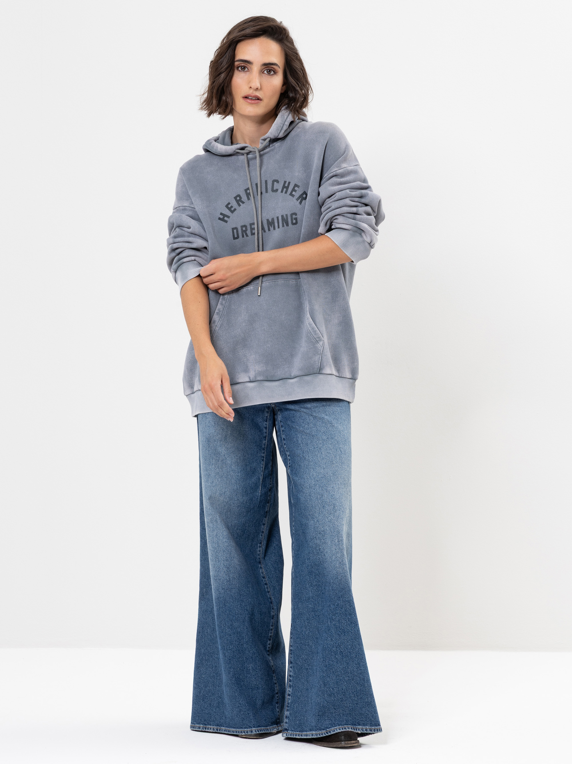 Eine Frau steht vor einem schlichten weißen Hintergrund und trägt einen lockeren grauen Kapuzenpulli mit Text und weit geschnittene Bluejeans. Sie hat kurzes braunes Haar und eine entspannte, lässige Pose, wobei eine Hand ihre Brust berührt.