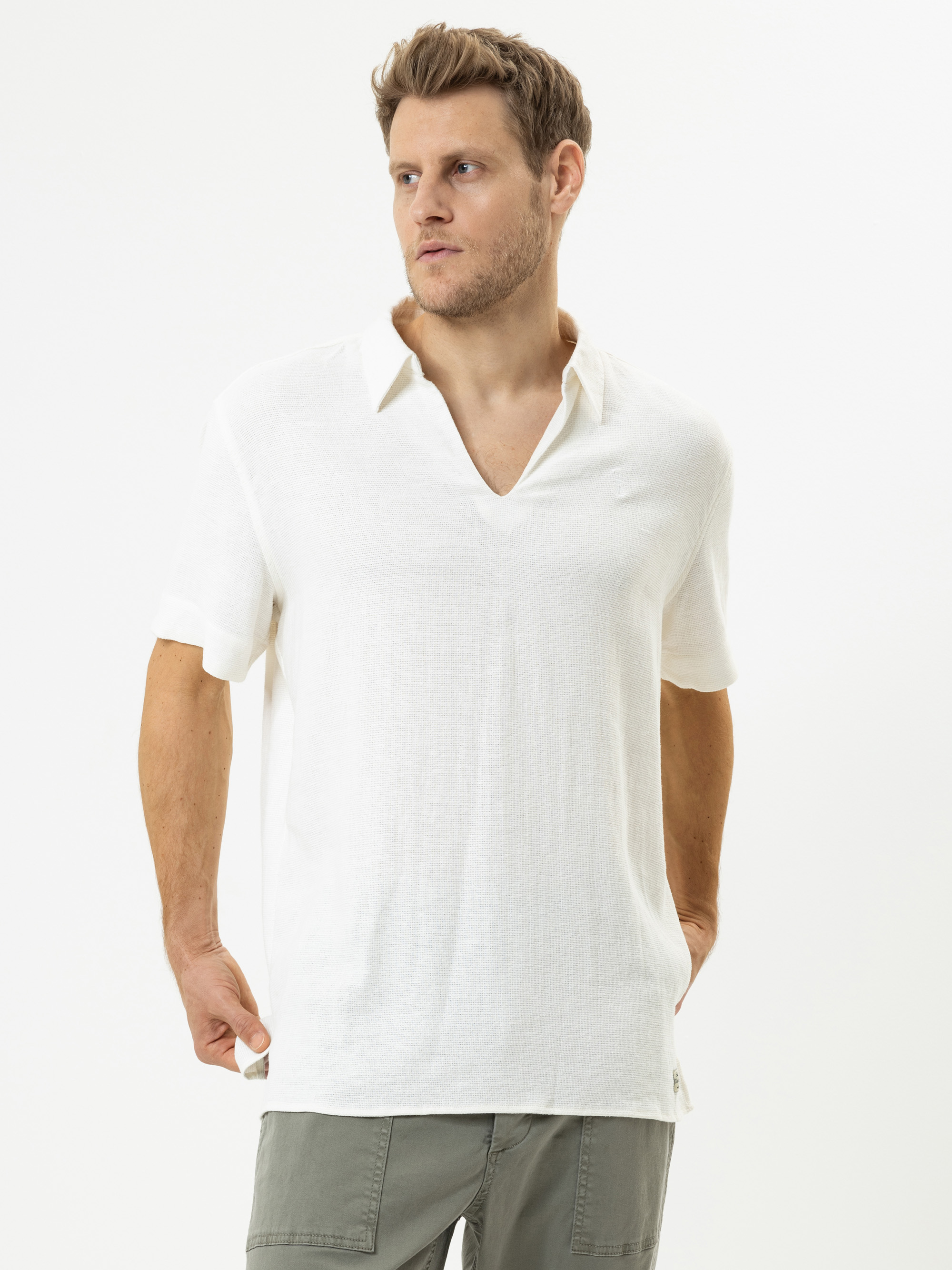 Ein Mann, der ein weißes, kurzärmeliges Hemd mit V-Ausschnitt und eine hellgrau-grüne Hose trägt, steht vor einem schlichten weißen Hintergrund und blickt leicht zur Seite, eine Hand in der Tasche.