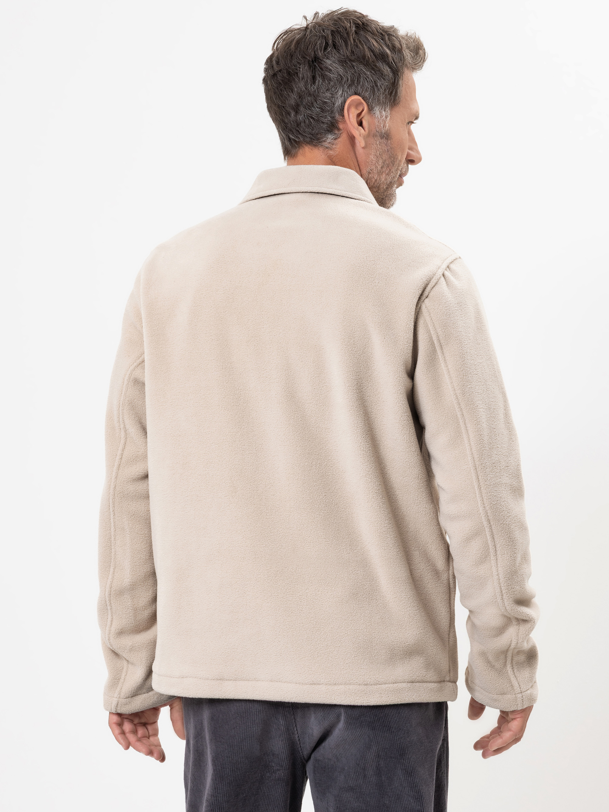 Ein Mann mit kurzen braunen Haaren, von hinten gesehen, trägt eine hellbeige Fleecejacke und eine dunkle Hose vor einem schlichten weißen Hintergrund.