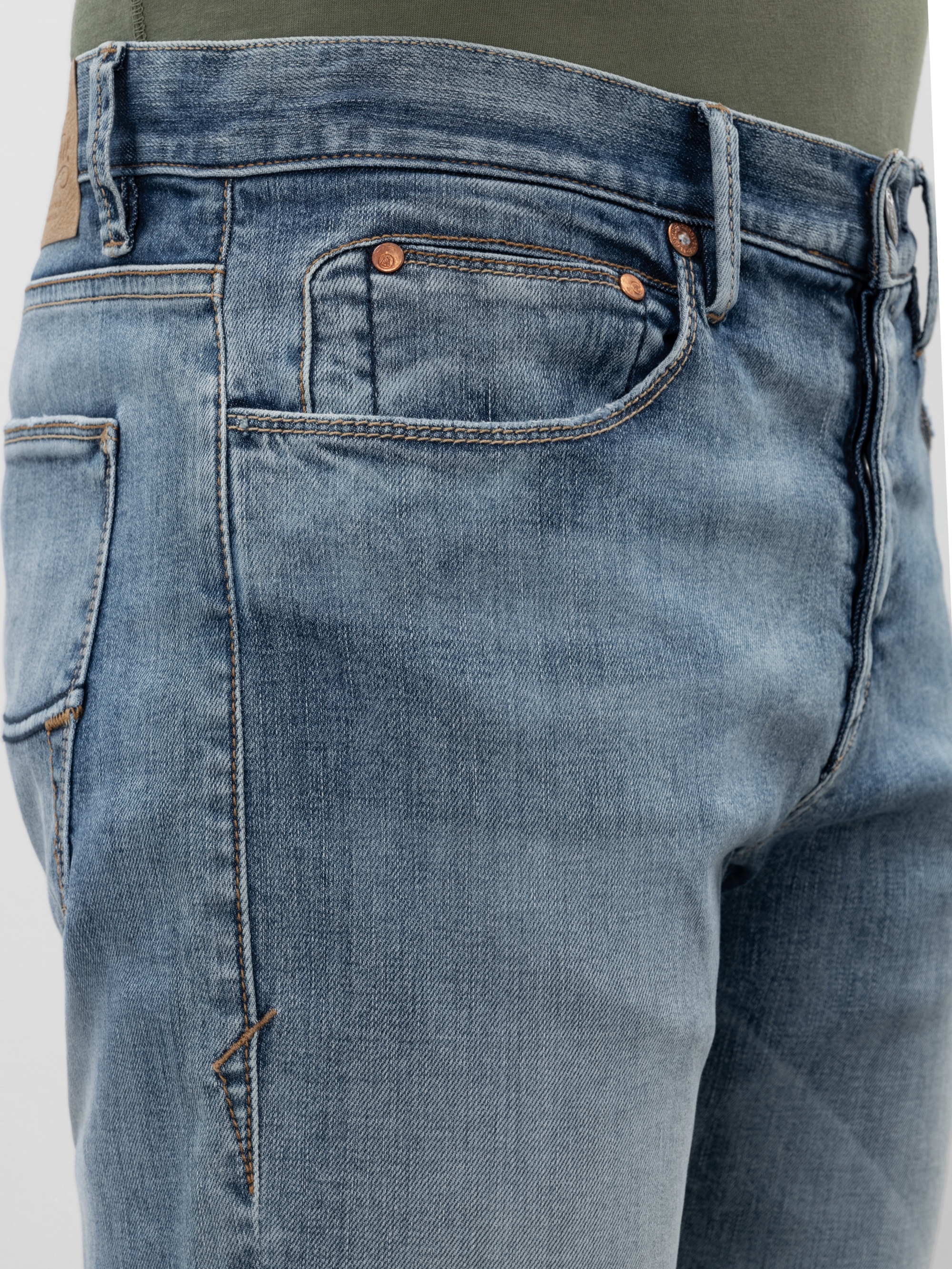 Nahaufnahme einer Person, die eine hellblaue Jeans mit kupfernen Nieten und sichtbaren Nähten trägt, dazu ein olivgrünes Hemd, das am Bund eingezogen ist.