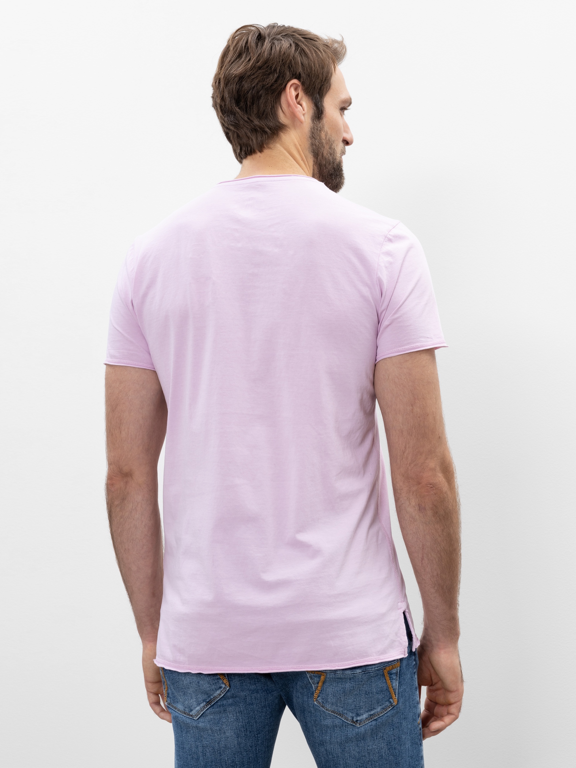 Ein Mann mit kurzen braunen Haaren steht mit dem Gesicht zur Kamera, trägt ein hellrosa T-Shirt und blaue Jeans vor einem schlichten weißen Hintergrund.