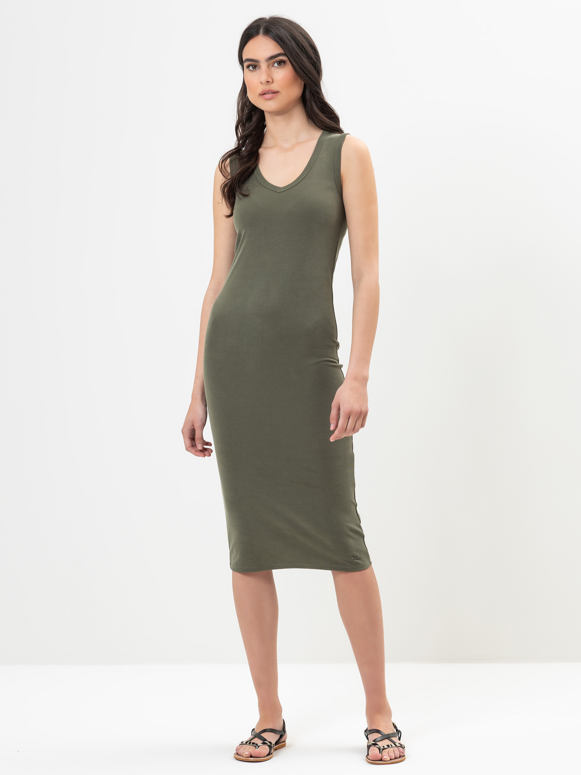 Eine Frau steht vor einem schlichten weißen Hintergrund und trägt ein olivgrünes, ärmelloses, knielanges, tailliertes Kleid und schwarze Sandalen. Sie hat langes dunkles Haar und einen neutralen Gesichtsausdruck.