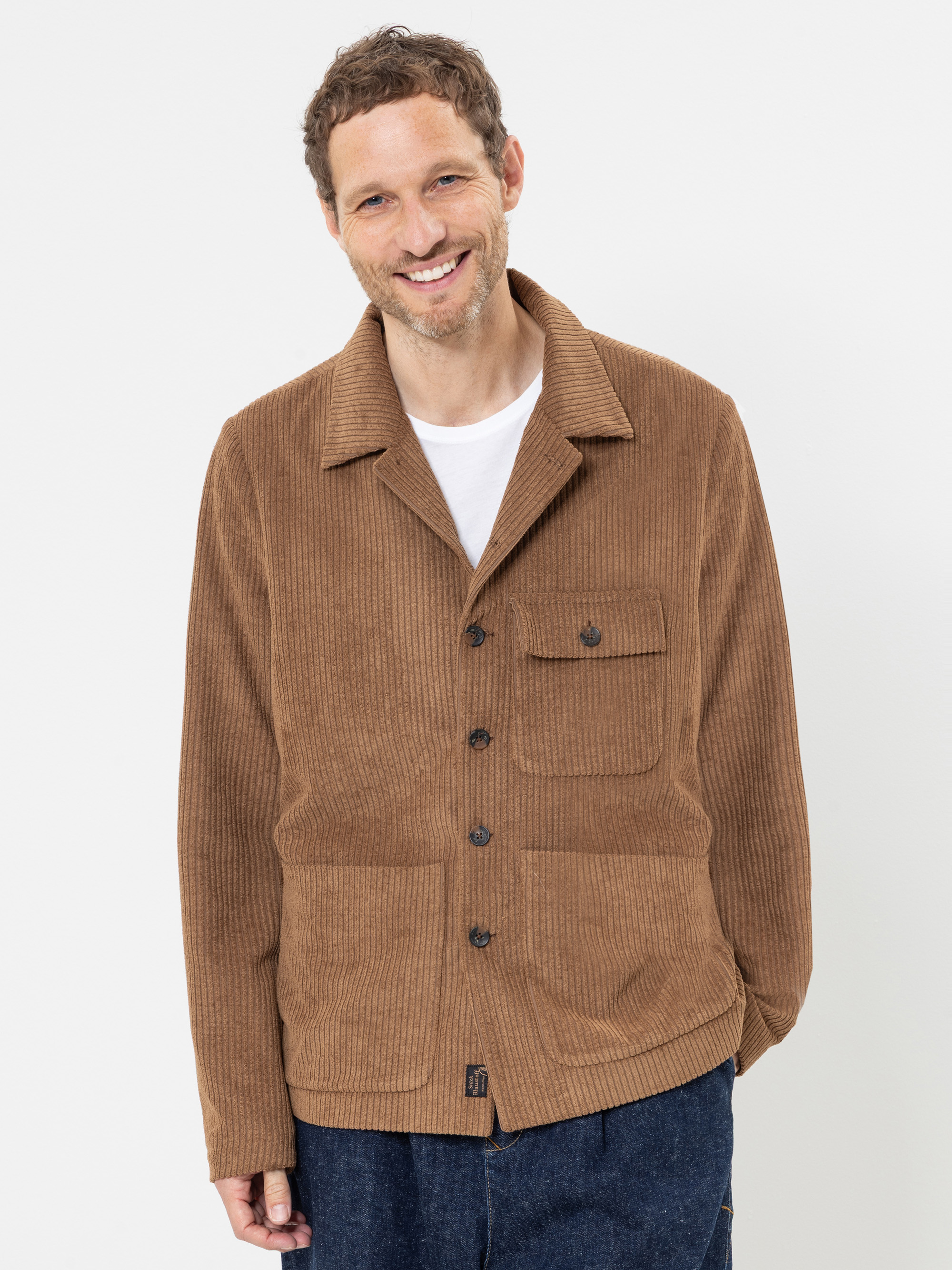Ein lächelnder Mann mit kurzen Locken, der eine hellbraune Cordjacke mit Knöpfen über einem weißen Hemd und einer dunkelblauen Hose trägt und vor einem schlichten weißen Hintergrund steht.