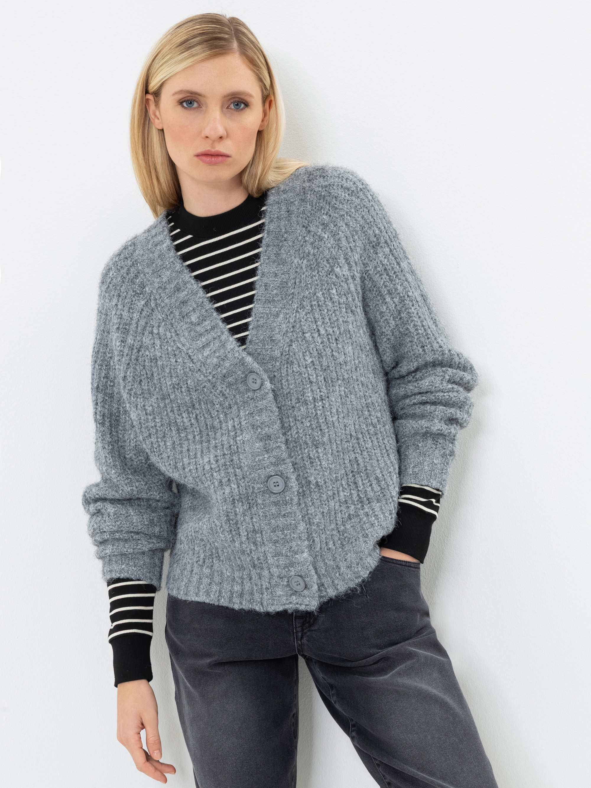 Eine Frau mit glattem blondem Haar trägt eine graue Strickjacke mit Knöpfen über einem schwarz-weiß gestreiften Shirt, dazu schwarze Jeans. Sie steht vor einem weißen Hintergrund und blickt mit ausdruckslosem Gesicht in die Kamera.