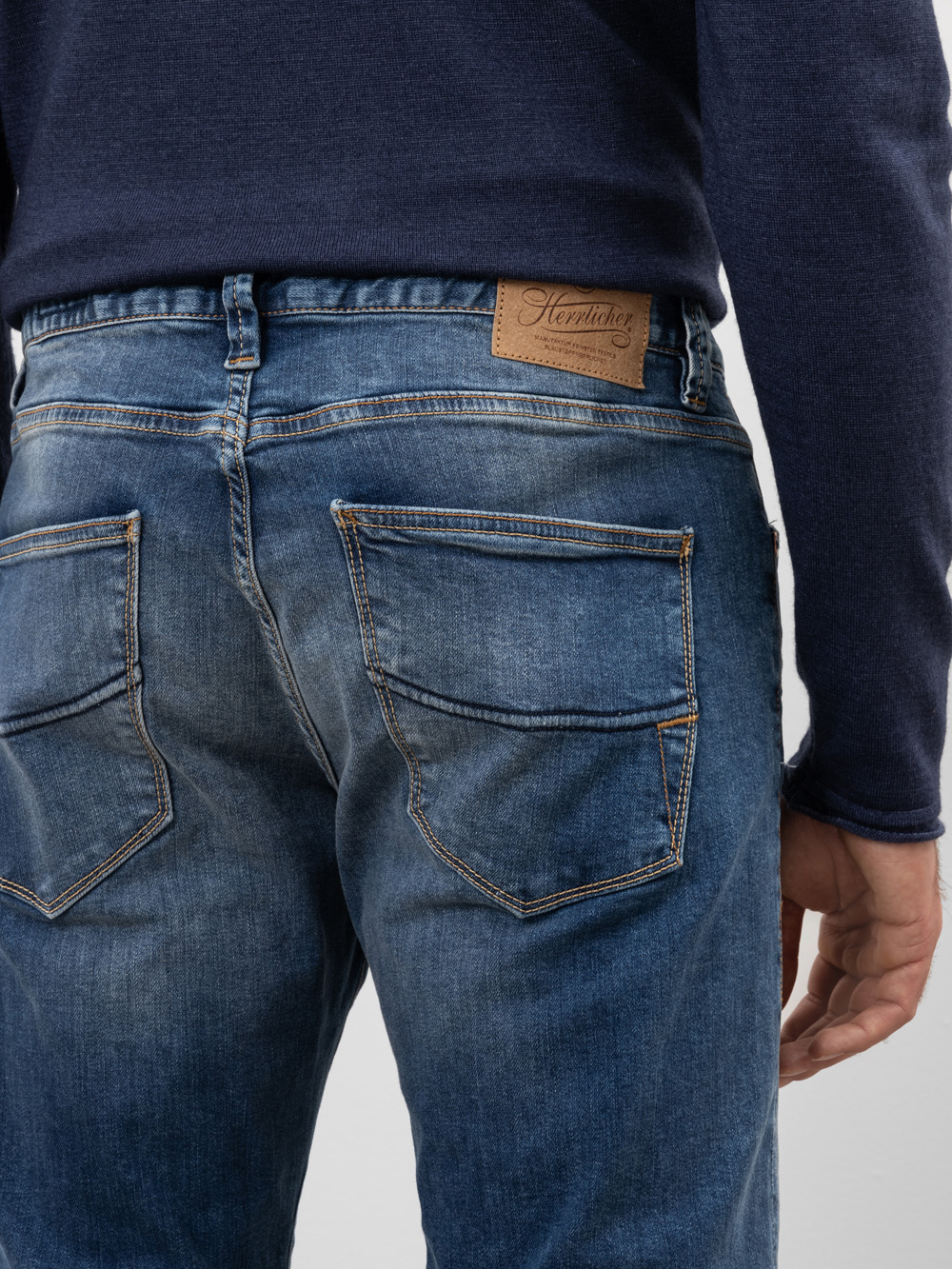 Eine Person, die eine blaue Jeans und einen marineblauen Pullover trägt, wird von hinten gezeigt. Die Jeans hat zwei Gesäßtaschen mit markanten Nähten und einen braunen Lederaufnäher am Bund.
