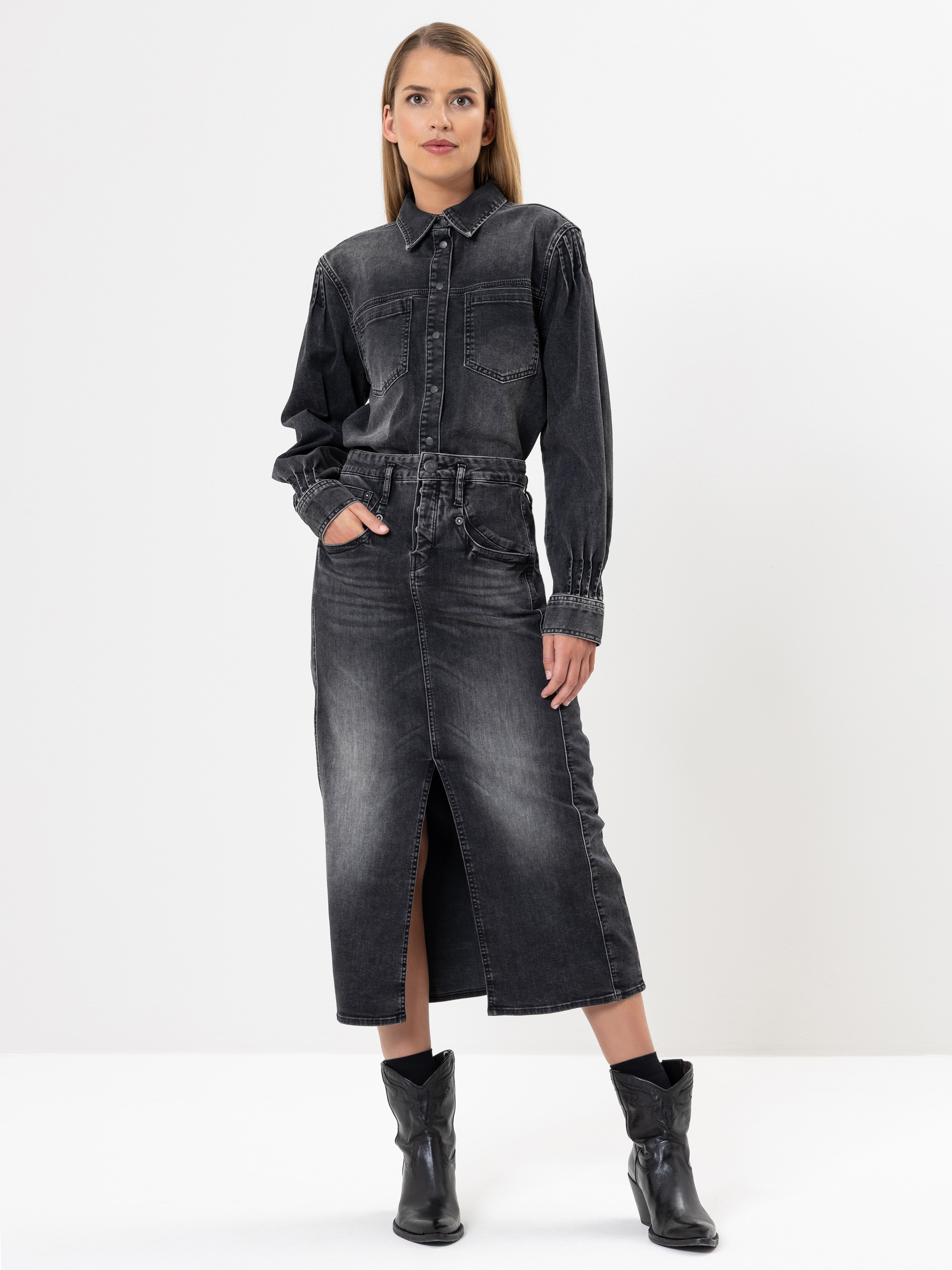 Eine Frau steht vor einem weißen Hintergrund und trägt ein schwarzes Jeanshemd und einen dazu passenden hochtaillierten Midirock mit einem Schlitz vorne. Sie hat eine Hand in der Tasche und trägt schwarze Ankle Boots.