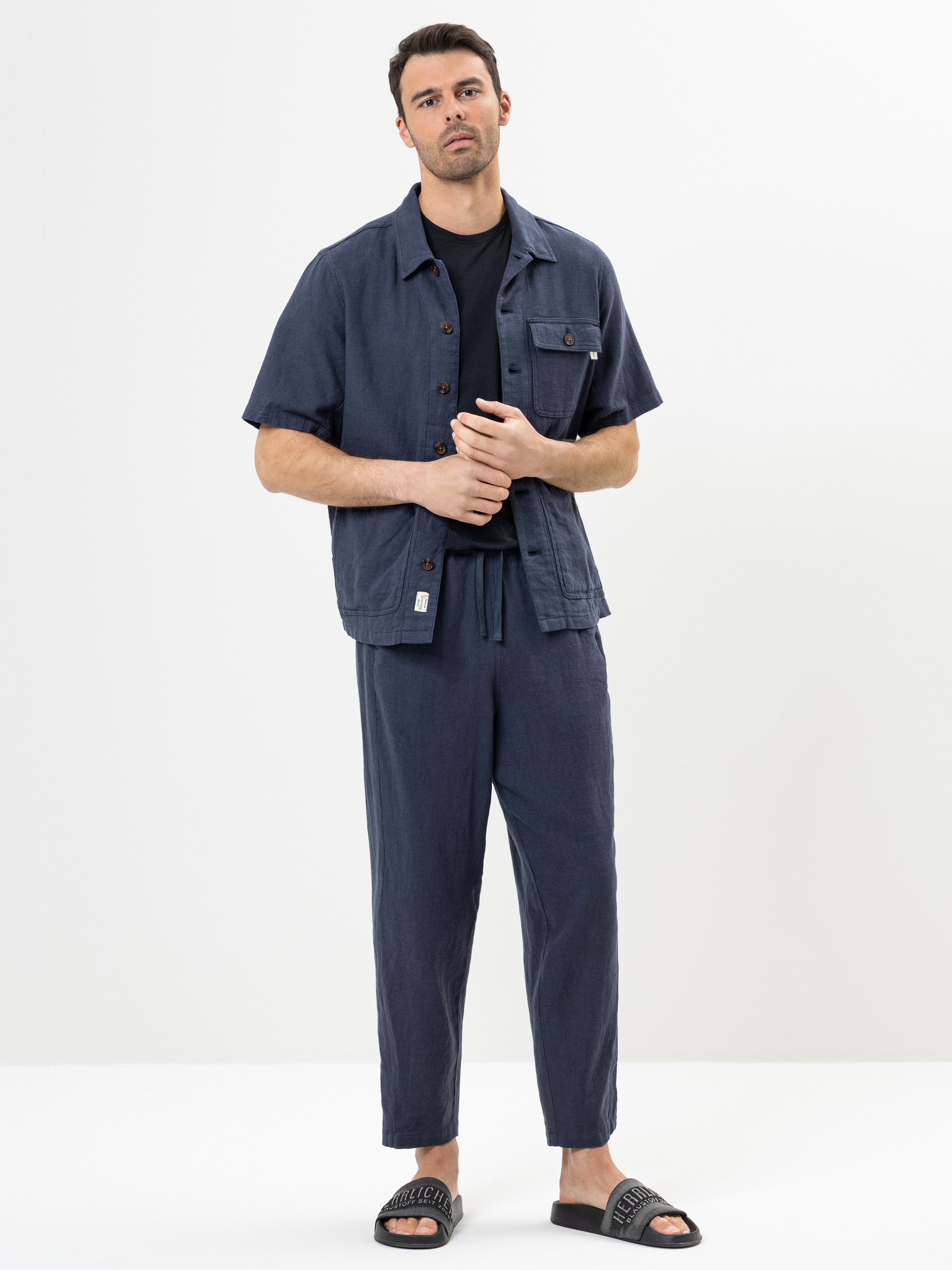 Ein Mann steht vor einem schlichten weißen Hintergrund und trägt ein marineblaues, kurzärmeliges Button-up-Hemd, eine marineblaue Hose, ein schwarzes T-Shirt und schwarze Slide-Sandalen. Er hat kurzes dunkles Haar und einen neutralen Gesichtsausdruck.