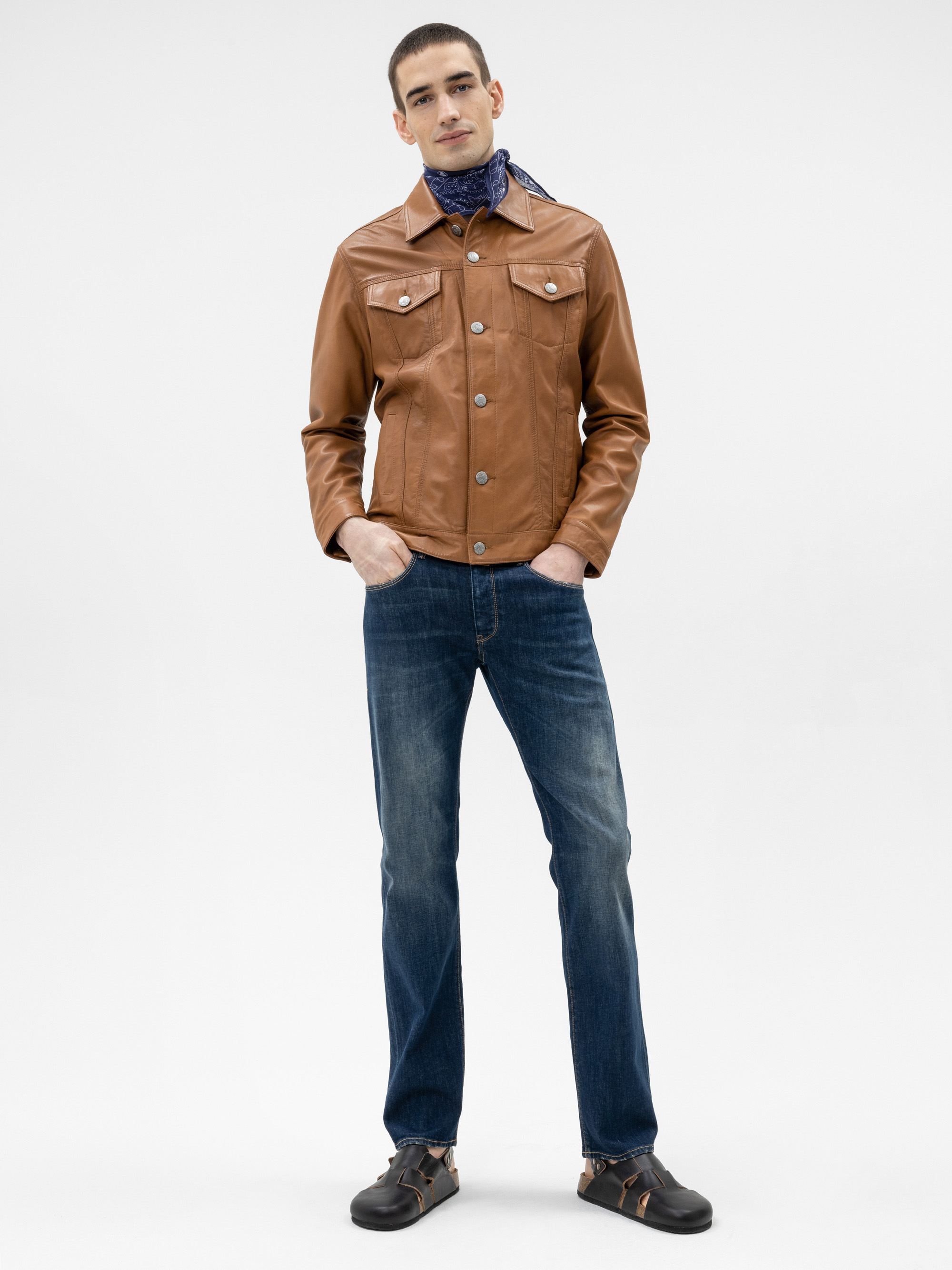 Ein Mann steht mit den Händen in den Taschen, trägt eine braune Lederjacke, blaue Jeans, schwarze Schuhe und einen blau gemusterten Schal vor einem schlichten weißen Hintergrund.